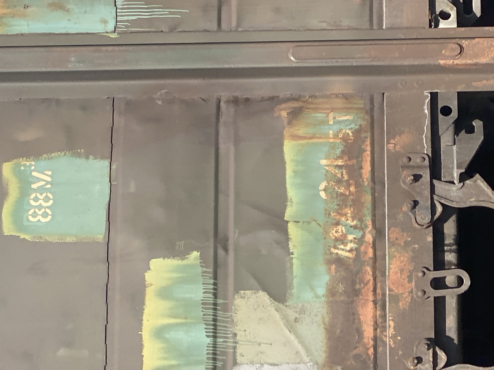 The image displays a close-up view of a rusty, dark-colored train car door. The surface is marked with faded, drippy paint in shades of green, yellow, and orange, suggesting weathering and age. Several stenciled numbers are visible: "883" in white on a light green rectangular patch at the top, and "4882457" in white on a greenish-blue patch towards the bottom, partially obscured by rust. The lower portion of the image shows metal hardware, likely part of the door's locking mechanism, also exhibiting signs of rust and wear. There is no discernible location within the city of Novokuznetsk, Russia, identifiable from this image. The scene is devoid of any people or visible actions.