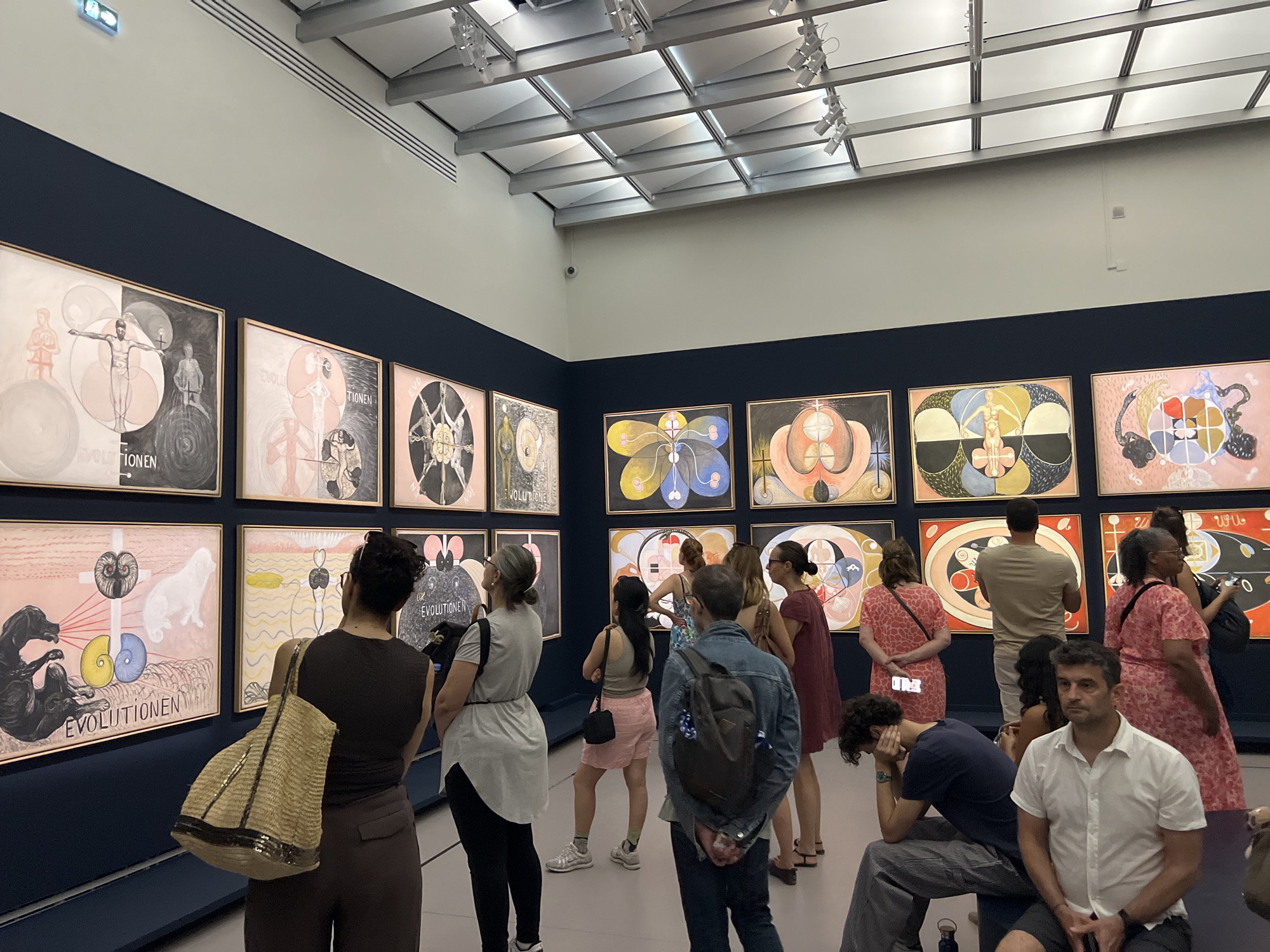 This is a medium shot of an art gallery exhibiting a series of paintings on a navy blue wall. The paintings appear to be part of a themed collection, possibly focusing on evolution, as indicated by the repeated word "Evolutionen" on some of the canvases. The artworks are framed and arranged in a grid-like fashion across two rows.
Several people are viewing the art. In the foreground, a man with dark curly hair is leaning forward, resting his head in his hands, wearing a navy blue t-shirt and striped pants. To his right, a man in a white collared shirt and dark shorts is seated and looking forward. Behind them, a woman with dark hair and glasses is standing, wearing a dark tank top and brown pants, carrying a woven straw bag.
Further into the gallery, other visitors are seen observing the paintings. A woman in a grey t-shirt and black pants is looking at a painting on the left. A woman in a light grey shirt and pink shorts is standing near her. More people are visible in the background, including a woman in a red patterned dress and another in a maroon dress.
The gallery space is well-lit, with a modern ceiling featuring a grid of lights. The walls are painted a light color above the navy blue exhibition wall. The floor appears to be a neutral color. The overall atmosphere is that of a public art exhibition.
The art style is figurative and symbolic, with recurring motifs like human figures, geometric shapes, and some animal imagery, particularly a black dog in one of the paintings. The color palette varies across the artworks, with some featuring muted tones and others bolder colors. The paintings are approximately eye-level for a standing adult. The man in the white shirt in the foreground is likely seated on a bench or low structure.