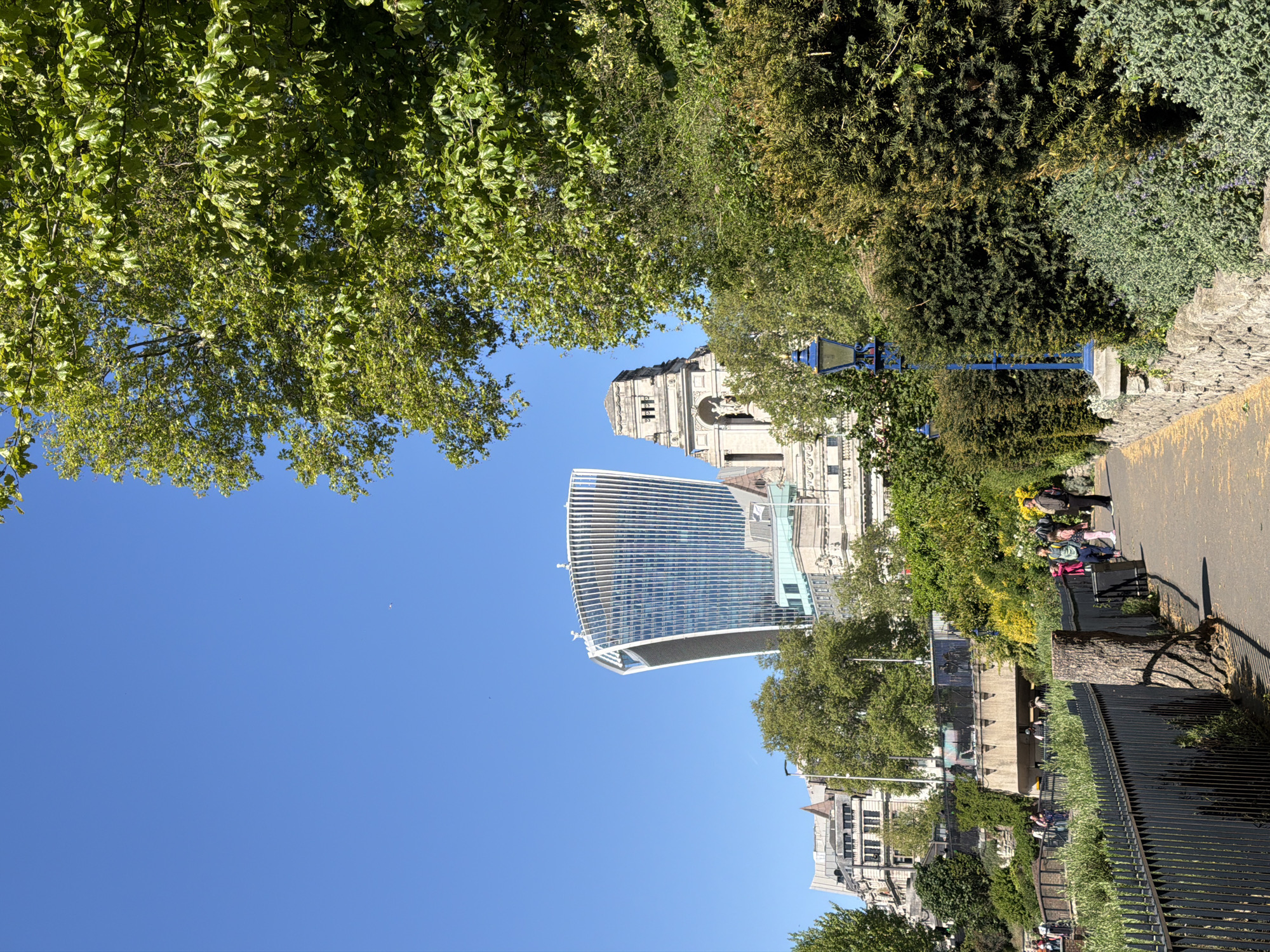 The image depicts a view in London, United Kingdom, showcasing the "Walkie Talkie" skyscraper (20 Fenchurch Street) juxtaposed with an older, classical building, possibly a church or civic structure. The scene is framed by foliage from trees overhead, with a clear blue sky in the background. A paved pathway runs along the right side of the image, bordered by a metal fence and greenery. Several people are visible along the pathway, some seated on a bench. A street lamp is positioned near the pathway. The image presents a contrast between modern architecture and historical buildings amidst an urban green space.