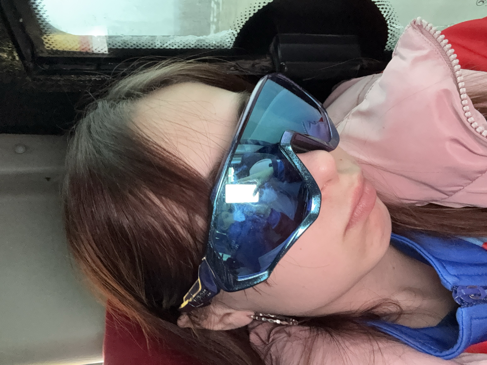 A young woman is lying down, possibly sleeping, with blue sunglasses covering her eyes. She appears to be in a vehicle, based on the window and vehicle interior visible. She is wearing a blue jacket with a zipper and a pink jacket is also visible. The image was taken in Челва, Russia.