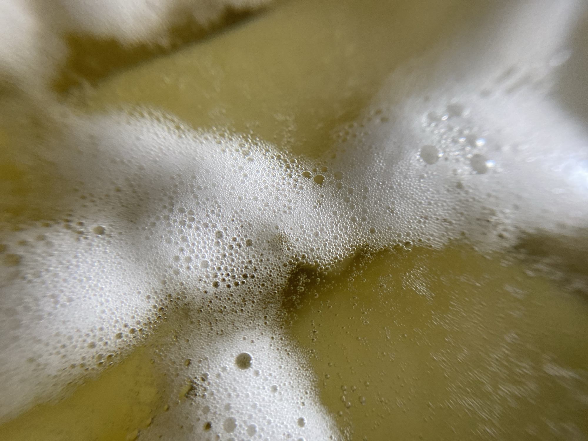 An extreme close-up shot shows a thick layer of white foam with many small bubbles covering the surface of a yellow liquid. The foam is uneven, with some areas appearing more dense and opaque than others. The bubbles vary in size, with some being almost microscopic and others more noticeable. The yellow liquid underneath the foam is visible through thinner sections, showing a light golden hue. The lighting highlights the texture of the foam, creating small glints and reflections on the bubbles. The image is abstract, focusing on the textural and visual qualities of the foam and liquid. The location is not determinable from the image.