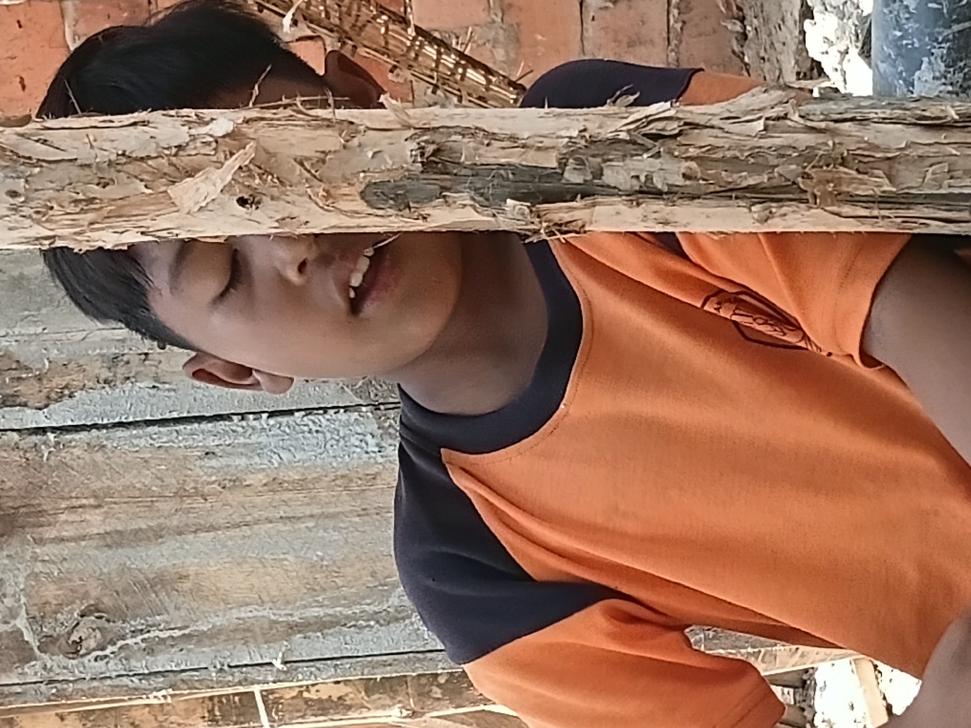 The image shows a close-up, angled shot of a young boy in Margo Rukun, Indonesia. He is wearing an orange t-shirt with dark blue sleeves. His eyes are closed, and his mouth is slightly open, suggesting he might be sleeping or resting. The boy is positioned behind a rough-hewn wooden plank, which is the dominant element in the foreground, obscuring part of his face and torso. The plank has a weathered, splintered texture.
Behind the boy, brickwork is visible, and parts of what appear to be thatched roofing or bamboo structures can be seen. The lighting suggests it is daytime, likely outdoors, given the natural textures and materials. The overall impression is one of a rustic, possibly impoverished, environment. There is no visible text in the image. The composition focuses on the boy's face and the textures of the wooden plank, creating a somewhat intimate and perhaps somber portrait.