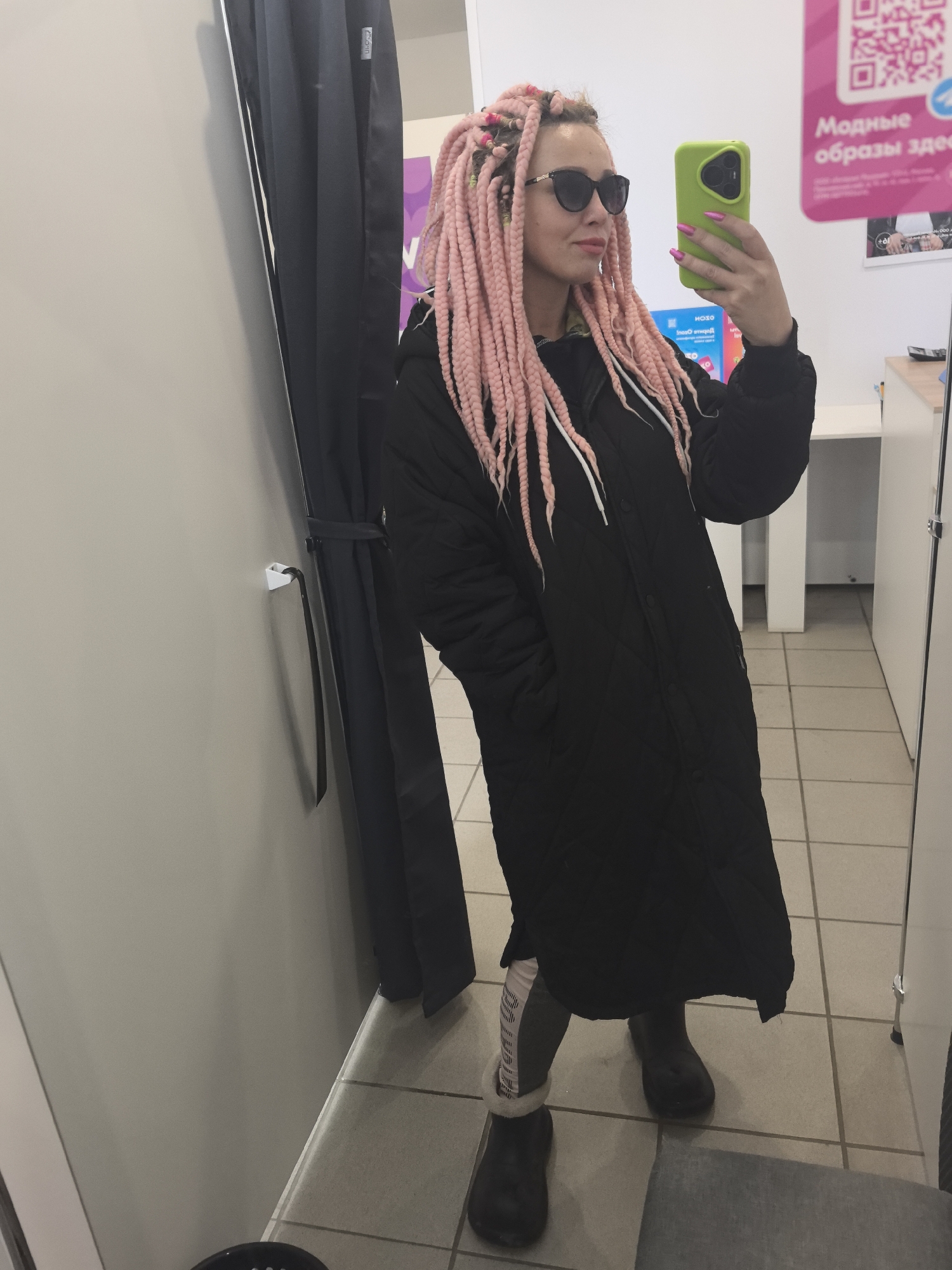 A woman is taking a selfie in a fitting room of a store. She is wearing a long black coat, dark sunglasses, and has pink braids. She is holding a green phone in her right hand. The fitting room has a gray wall, a curtain, and tiled floors. A poster is visible on the wall with Russian text "Модные образы здесь" (Fashionable looks here). The presence of the Russian text indicates the location is likely in Russia, but there is insufficient information to identify the city as Noviy.
