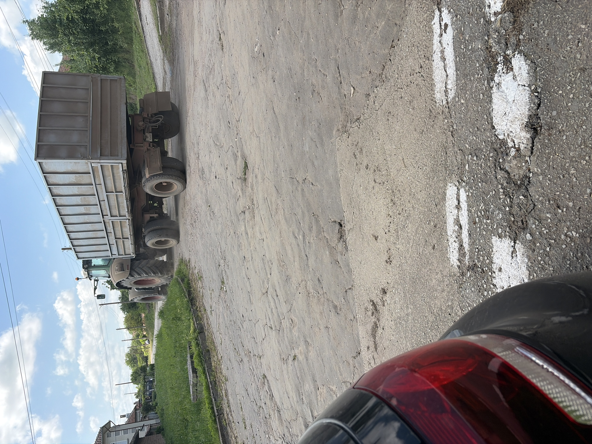 The image shows a tractor with a trailer driving on a cracked asphalt road in Kozlovets, Bulgaria. A car with a red taillight is partially visible in the lower right corner of the frame. The road has partially obscured white lane markings. Vegetation borders the road. Buildings and trees are visible in the background. The sky is visible with clouds.