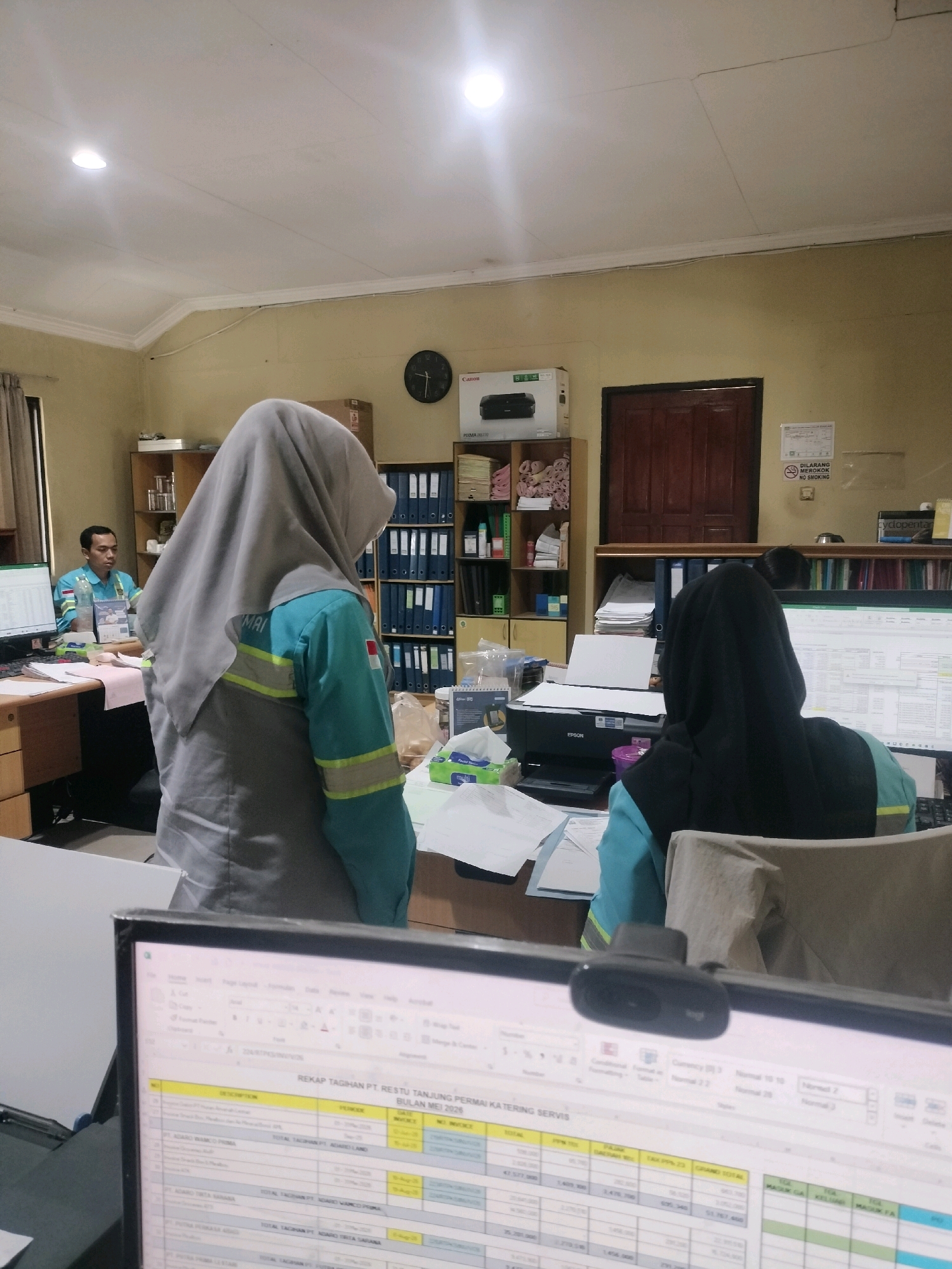In a brightly lit office, two individuals wearing teal and grey work shirts and head coverings are visible. One person, facing away from the camera, is seated in front of a computer monitor displaying a spreadsheet. A webcam is positioned on top of the monitor. To the left, another person, wearing a teal shirt, is seated at a desk, also facing a computer monitor. The office environment includes shelves filled with binders and files, a printer, and papers scattered across desks. A clock on the wall indicates the time is approximately 12:26. The office appears to be located in Maburai, Indonesia, as suggested by the presence of Indonesian language text on a sign which translates to "No Smoking".