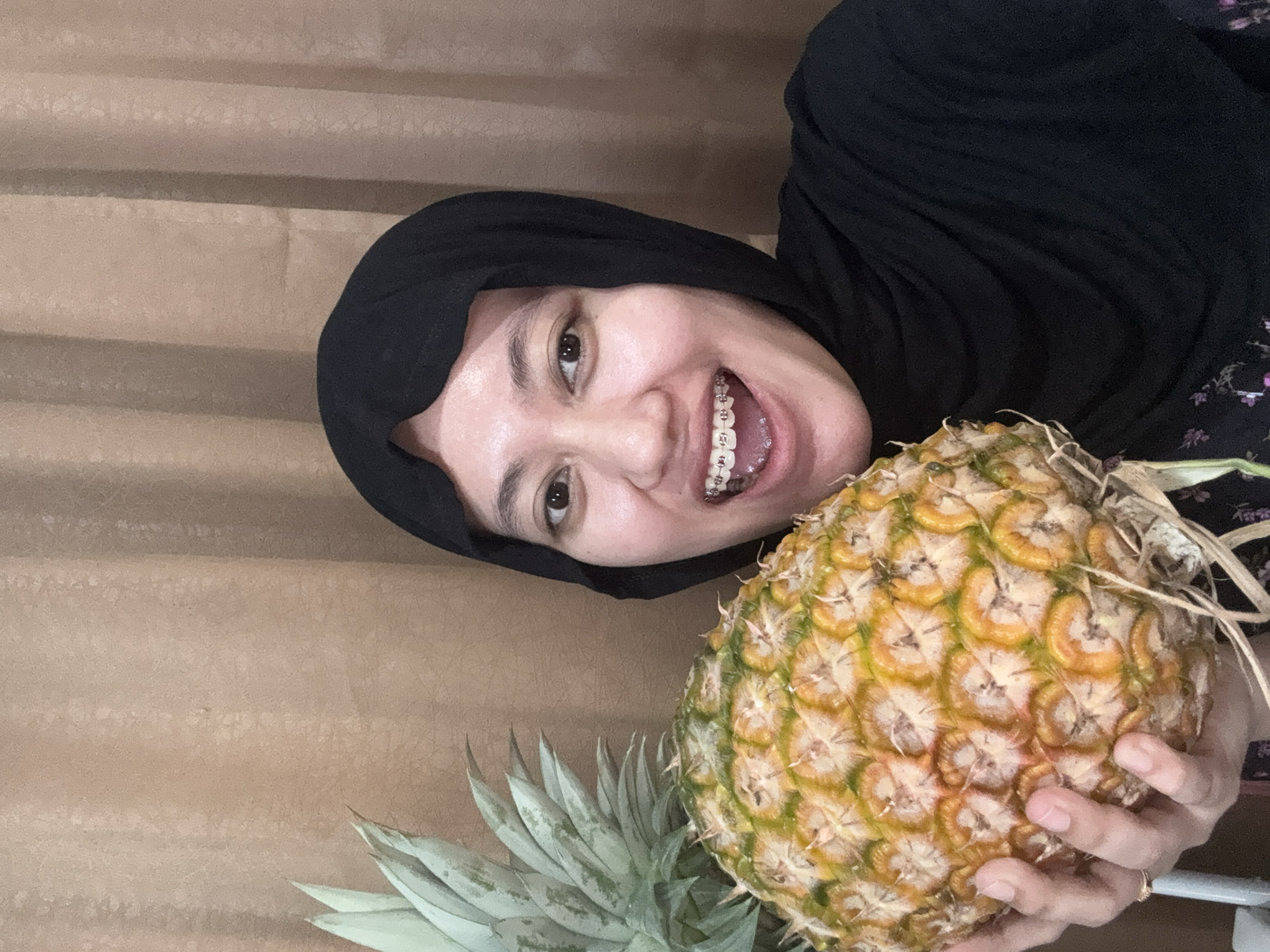 A woman wearing a black hijab is holding a pineapple. She has dental braces. The background appears to be a beige curtain. The location is Gadingrejo, Indonesia.