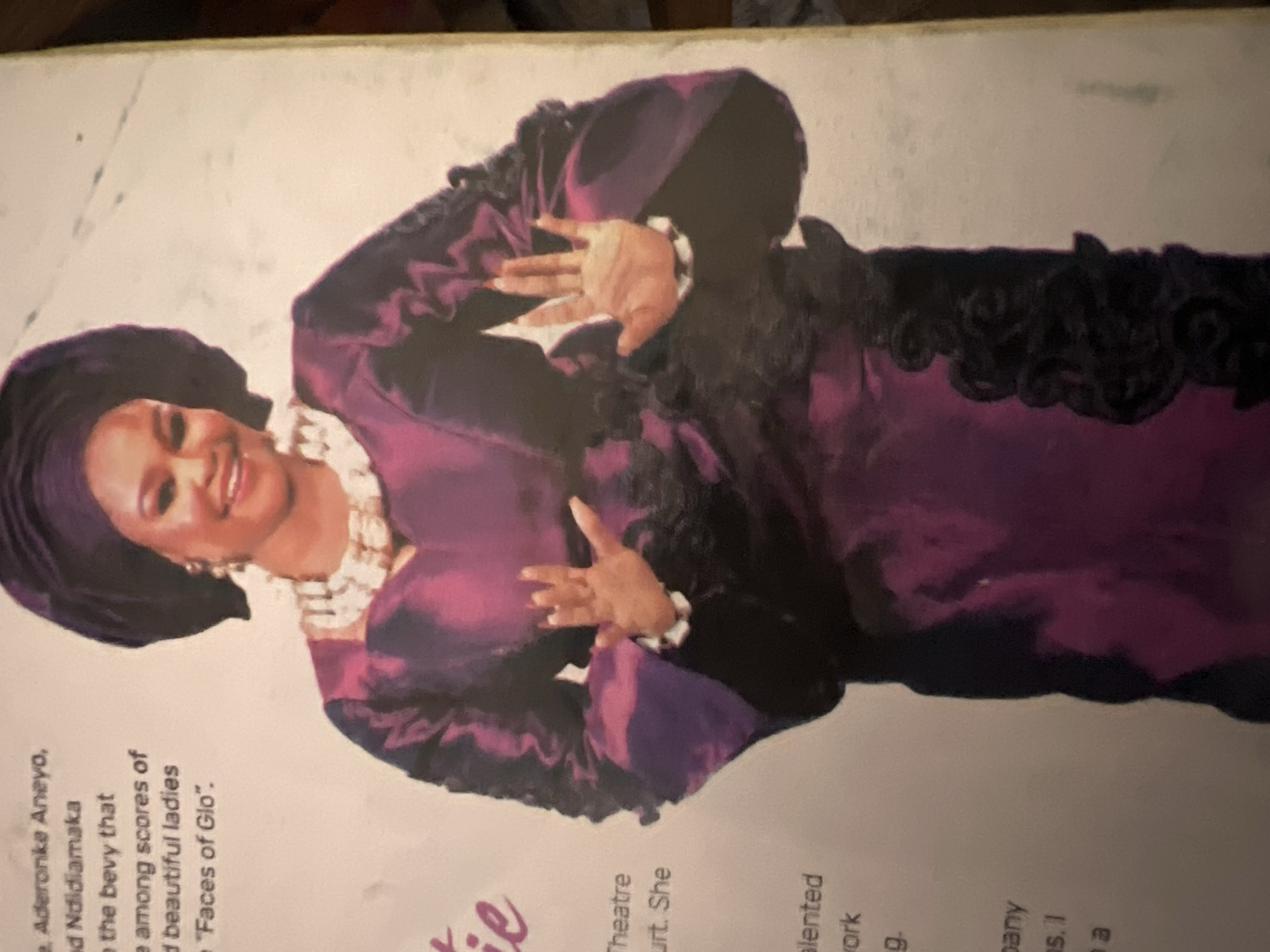 A portrait of a woman dressed in traditional Nigerian attire, specifically a purple iro and buba with a matching headwrap, is displayed. She wears a pearl necklace and large earrings. Her hands are raised, with palms facing forward, as if in a gesture of greeting or explanation. The background is a plain, light-colored surface, likely a wall or a printed page, with some text visible to the left of the woman. The text appears to be biographical information, possibly relating to her career or an event. There is no information in the image to suggest the location is in Jalingo, Nigeria.