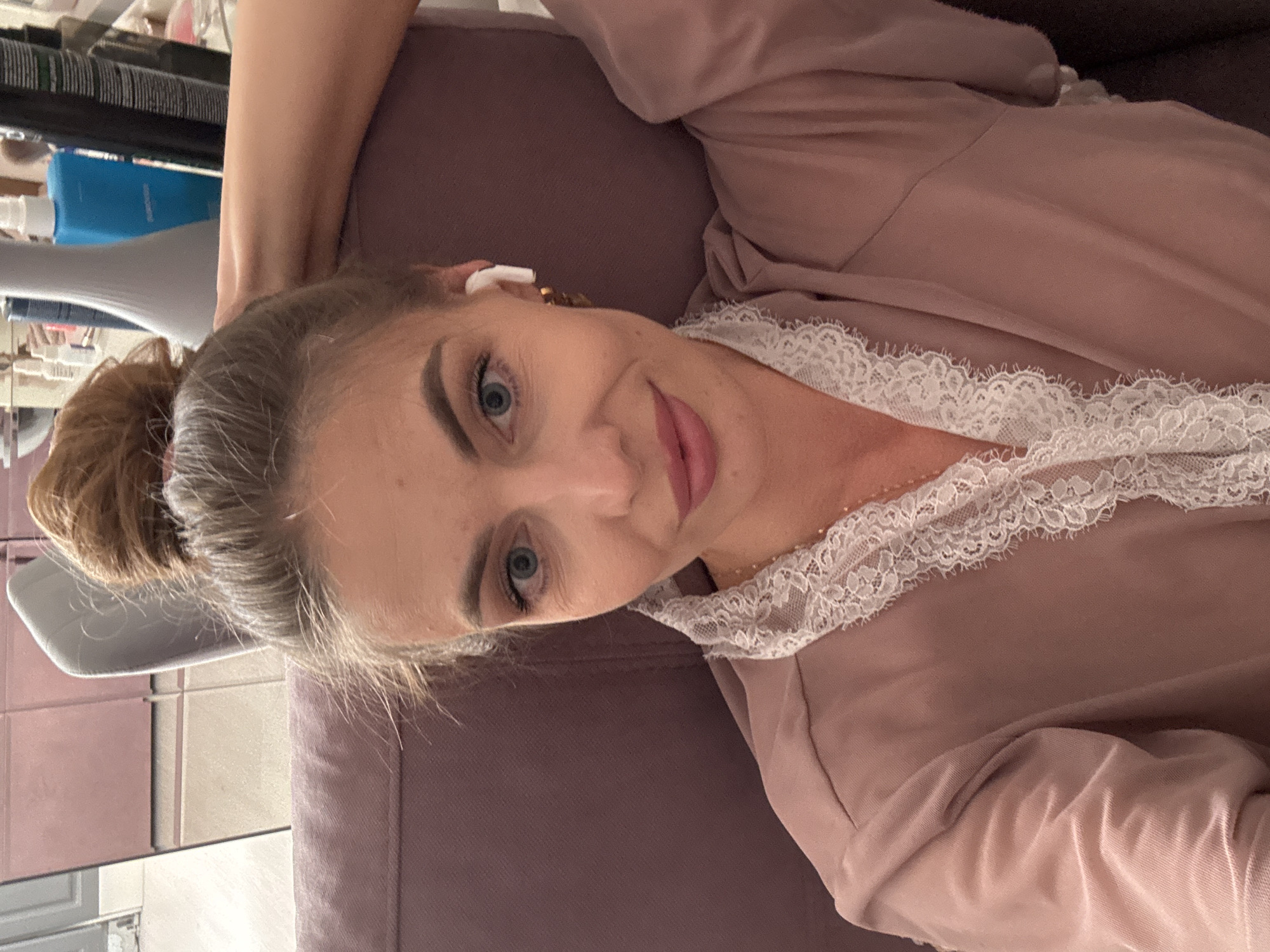 A woman with blonde hair in a bun is sitting on a pink sofa, looking at the camera. She is wearing a pink robe with white lace trim and white wireless earbuds. Her left arm is resting on the back of the sofa, with her hand behind her head. The background shows a shelf with various items, including bottles and containers, suggesting a dressing area or vanity. The overall scene is indoors, likely in a residential setting, and does not contain any information about the city of Sochi, Russia.