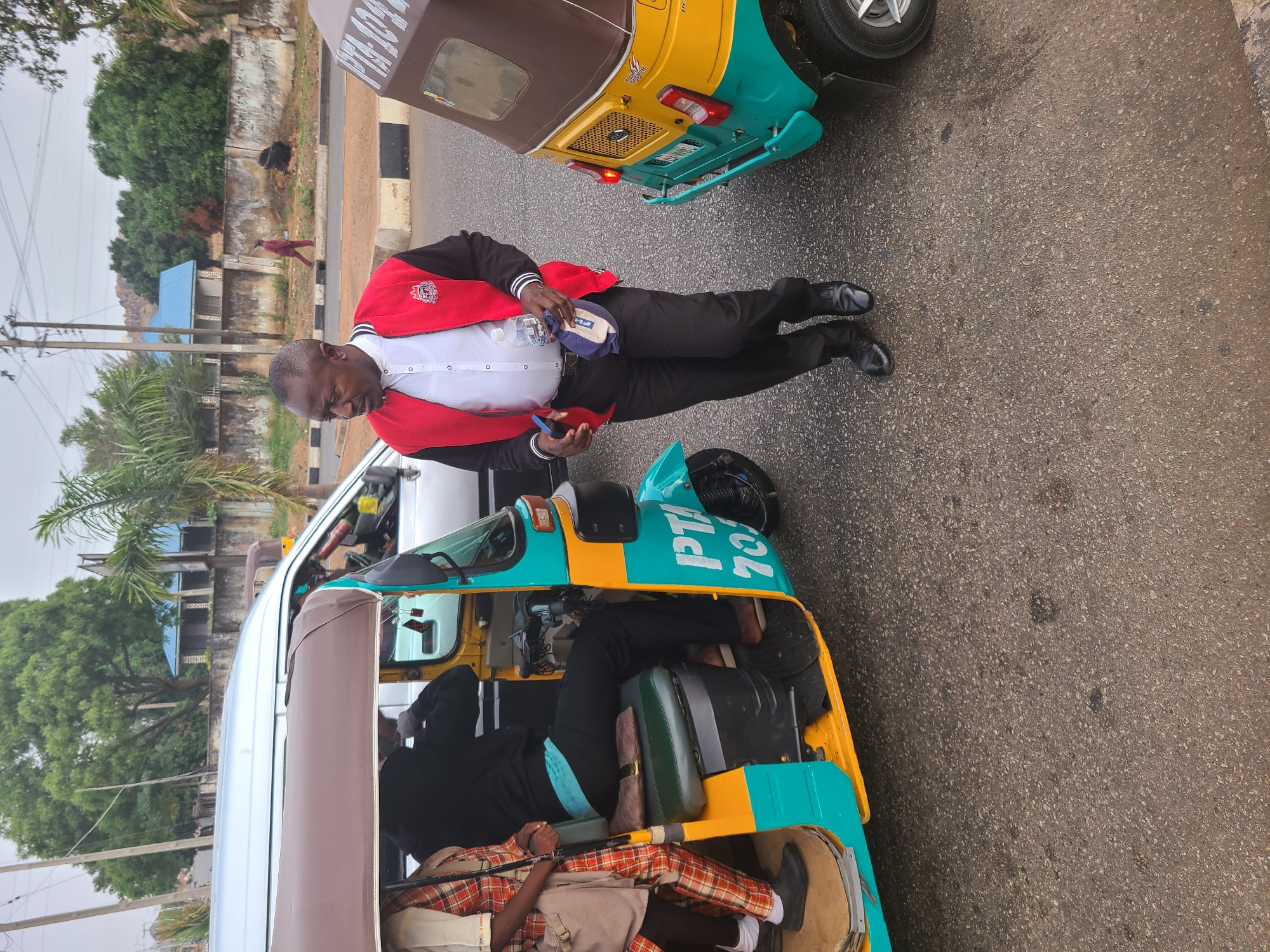 A man in a red and black jacket, white shirt, and black pants stands on a grey asphalt road in Jos, Nigeria. He is positioned between two light blue and yellow auto rickshaws (tuk-tuks). He holds objects in his hands, appearing to look at them. One tuk-tuk has two visible passengers inside. The environment includes buildings, trees, and a road. A person walks on the road in the background.