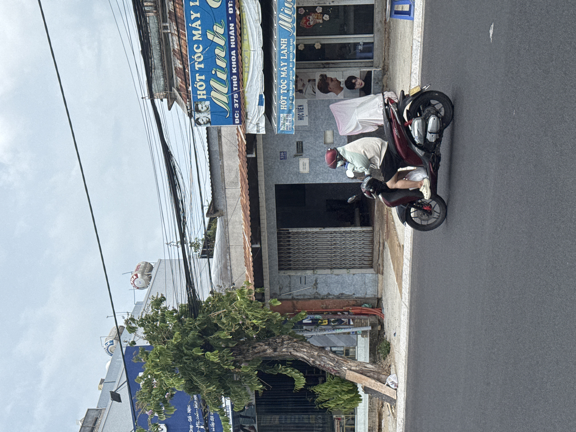 The image depicts a street scene in Phan Thiết, Vietnam, on an overcast day. A person wearing a light-colored hoodie, dark shorts, a helmet, and white Crocs is riding a dark red scooter on the right side of the asphalt road. The scooter has chrome accents and a yellow license plate.
In the background, there are several buildings. The closest building on the right has a blue awning with Vietnamese text and images of men's hairstyles, indicating a barbershop or hair salon. A blue plastic stool is visible on the sidewalk. Adjacent to this, to the left, is a building with a gray facade and a large metal roll-up door. To the left of that, a tree with lush green foliage leans over the sidewalk, supported by wooden planks. Further left, a building with a blue facade displays text in Vietnamese. On the roofs of some buildings, large water tanks are visible. Power lines are strung across the sky, creating a dense network above the street. The overall environment suggests a commercial area in Vietnam.