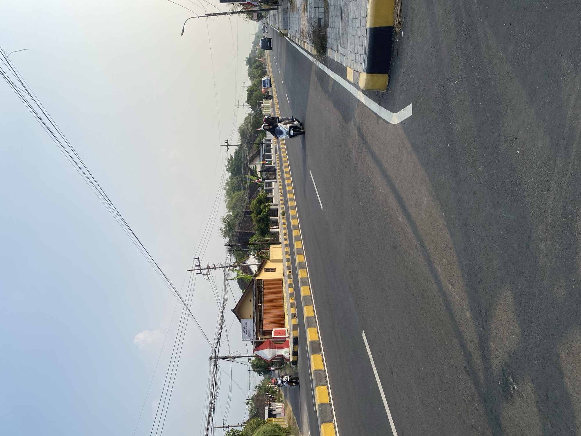 The image depicts a straight, paved road in Sugihwaras, Indonesia, under a clear, pale blue sky. The road has a white dashed line dividing the lanes and a white solid line near the curb. The sides of the road are lined with a curb painted with alternating black and yellow blocks. A sidewalk paved with square stones runs along the right side of the road.
In the foreground, the asphalt road surface is visible with shadows cast from overhead objects, likely utility wires. In the middle ground, several vehicles are present. On the left side of the road, a person rides a white scooter. Further down the road, on the right lane, two people ride a white scooter, with the passenger in the back wearing a red helmet. In the distance, on the right side of the road, a black SUV and a white van are driving.
Along the left side of the road, there are buildings with yellow walls and orange-brown doors. One building has a sign that reads "NOTARIS CAHYANI, S.H., M.H.". Red and white flags, possibly Indonesian flags, are displayed on poles near the buildings. Trees and vegetation are visible beyond the buildings. On the right side of the road, a street lamp is visible. The overall scene suggests a typical day on a suburban or rural road in Indonesia.