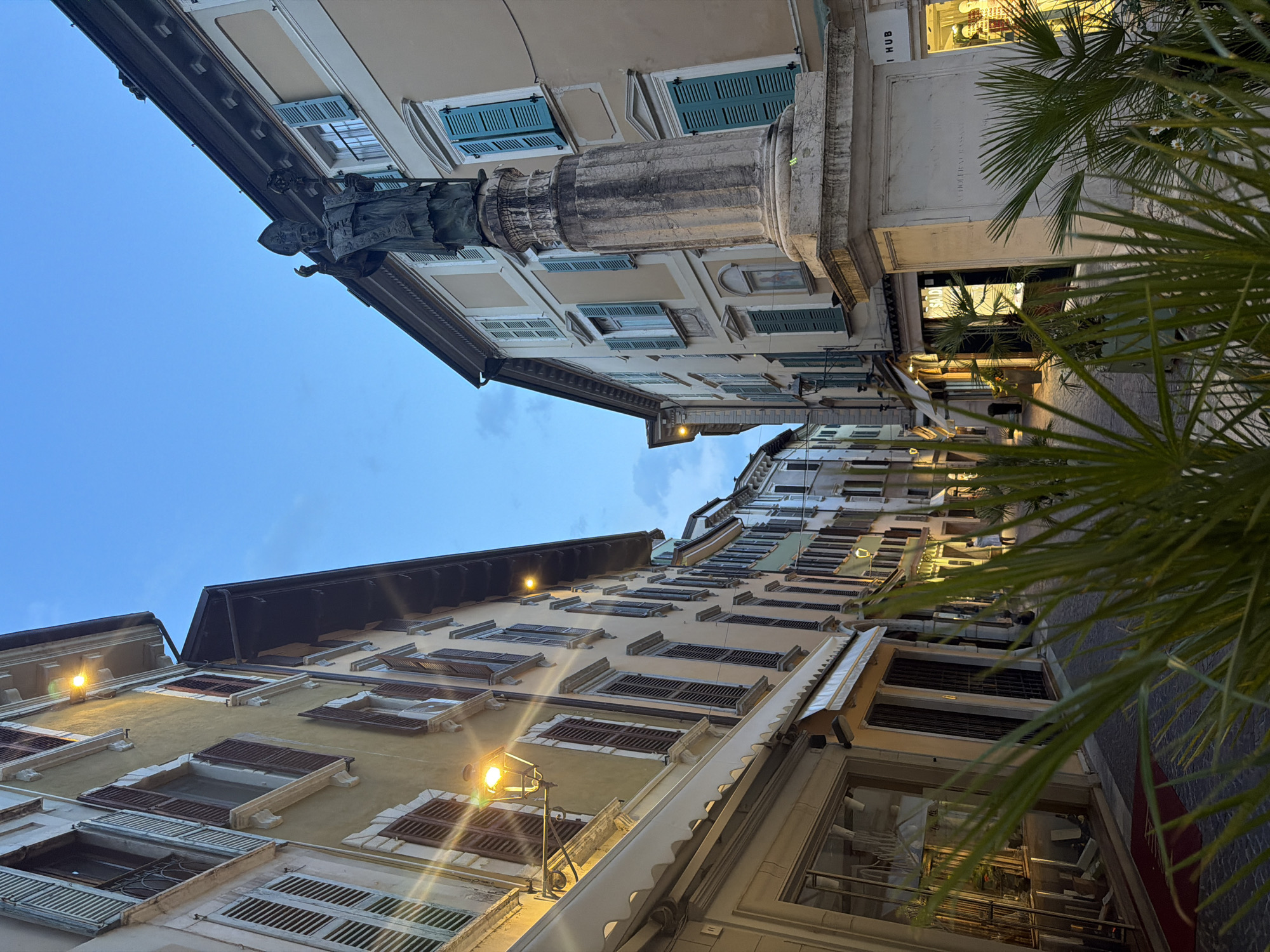 A vertical, low-angle outdoor photograph captures a street scene in Salò, Italy, as dusk settles. The sky above is a deep, muted blue with hints of clouds. Flanking the cobblestone street are tall, narrow buildings with cream and ochre facades, adorned with numerous windows featuring dark wooden shutters. Ornate street lamps with a warm yellow glow are mounted on the walls, casting pools of light.

In the foreground, on the right side of the street, a large stone pedestal supports a dark bronze statue of a robed figure wearing a miter and holding a staff, its arms raised. The statue faces towards the left side of the frame. In front of the pedestal and extending into the lower right corner of the image are large, green palm fronds, providing a natural element to the urban setting.

The street continues into the distance, lined with more buildings and shops. Some shop windows are illuminated, reflecting the lights from within and from the street. A few individuals are visible further down the street, one person appearing to walk away from the camera. The overall atmosphere is tranquil, with the softening light of early evening and the warm glow of the streetlights.