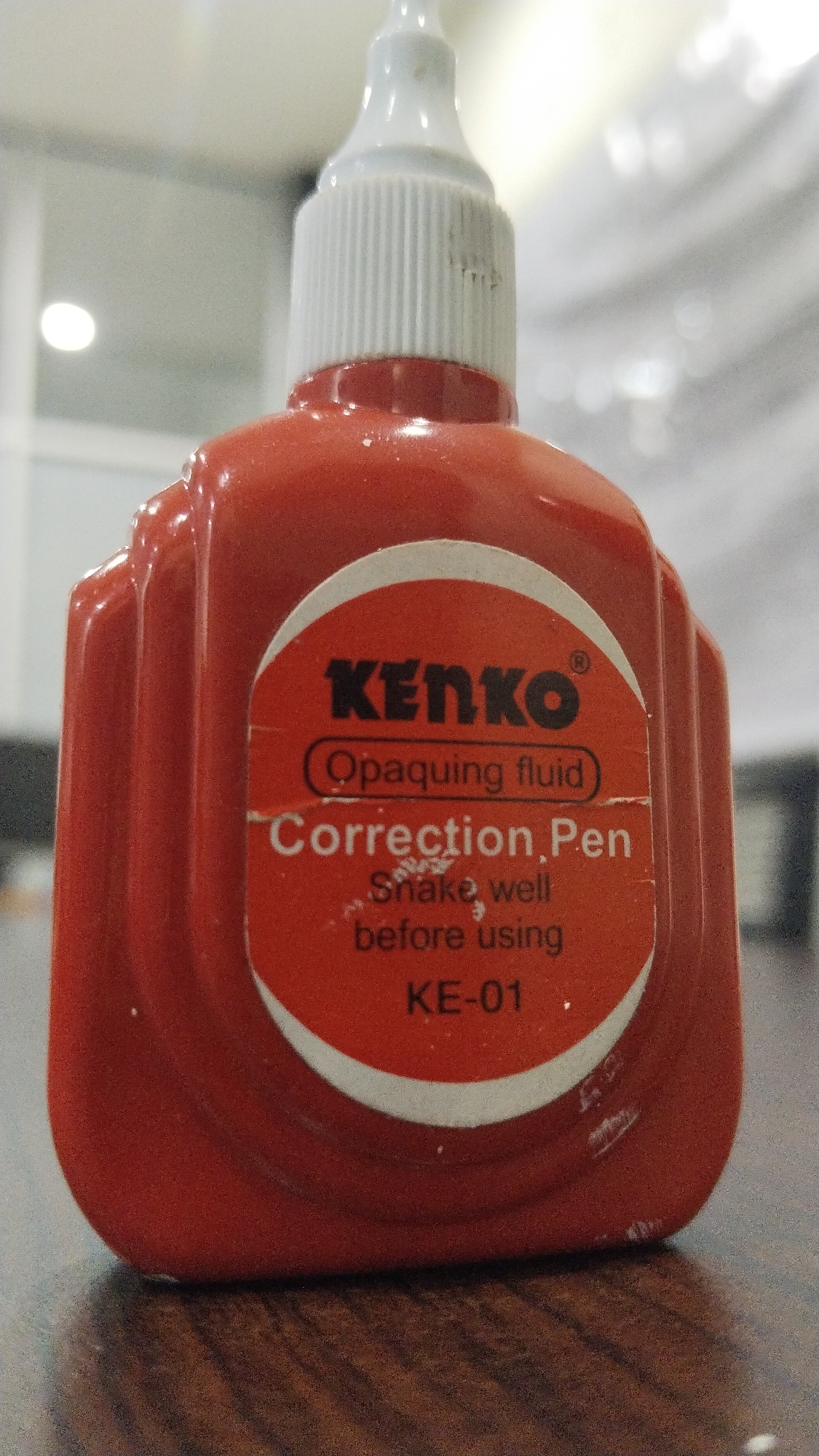 A close-up, eye-level, slightly low-angle, indoor shot shows a bright red bottle of "Kenko" correction fluid with a white cap. The bottle is positioned on a dark, wooden surface with a visible wood grain pattern. The background is blurred and out of focus, with some lighter areas suggesting indoor lighting and possibly office furniture or walls. A circular orange label is affixed to the front of the bottle, featuring black text. The label reads "KENKO" prominently at the top, followed by "Opaquing fluid" enclosed in an oval. Below this, it states "Correction Pen" and smaller text indicates "Shake well before using," and finally "KE-01." The lighting is somewhat diffuse, creating soft shadows and highlighting the texture of the bottle and the wooden surface. The image lacks any signs of human presence or activity, and the overall scene appears static and unpopulated. The location is identified as Tanjung Batu Kota, Indonesia, though there are no visual cues in the image to confirm this specific geographical placement.