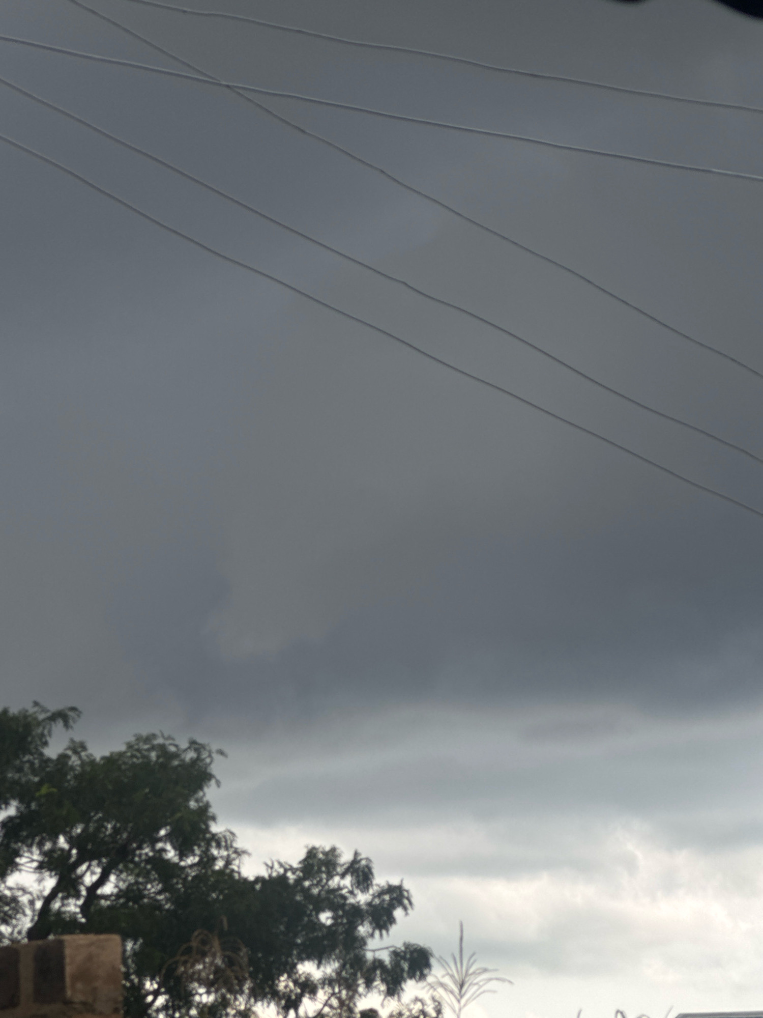 The image depicts a cloudy sky with several power lines crisscrossing it. In the foreground, there is a portion of a brick structure on the left side, partially obscured by foliage from a tree. The tree, with its dense green leaves, is a prominent feature in the lower left and center of the frame. The lower right portion of the image shows a faint glimpse of what appears to be part of a building or structure. The overall atmosphere is somber, with dark, heavy clouds suggesting an approaching storm or inclement weather.