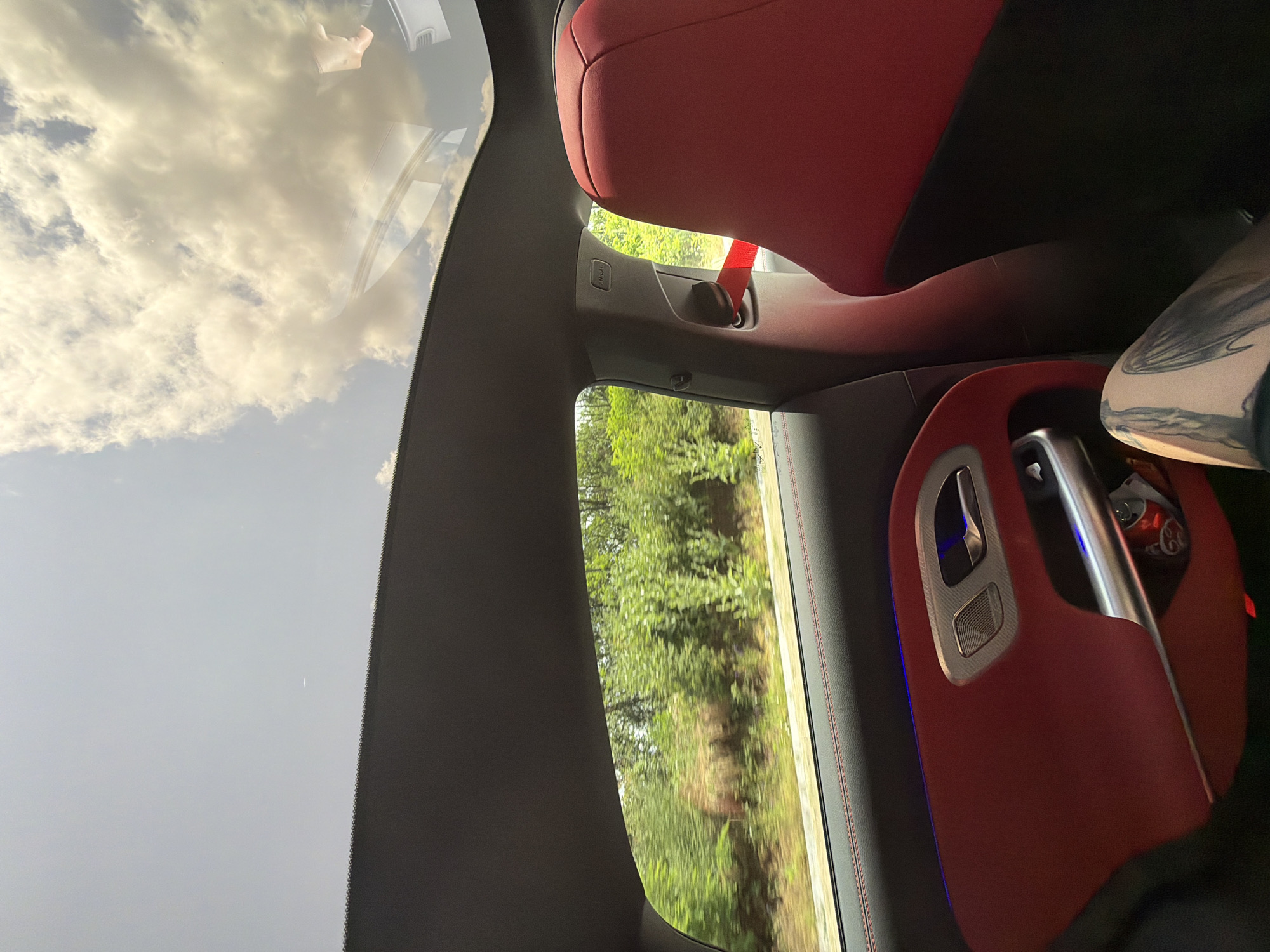 This image captures an interior view from a moving car, looking outwards and upwards. The car's cabin features prominent red seats and dark gray trim accented with red stitching, visible on elements like the door panel. A red seatbelt is seen, and a person's tattooed limb is partially visible on the right. A red-topped Coca-Cola can rests nearby. The door panel includes a silver handle, a speaker grille, and blue ambient lighting.
Through the side window, a blur of lush green trees and foliage indicates the vehicle is in motion. Above, the sky, viewed through the windshield or sunroof, shows bright white clouds against a light blue backdrop, suggesting daytime.
Visible text includes "AIRBAG" on a pillar cover and "Coca-Cola" on the can. The scene captures a moment of travel in Sadovets, Bulgaria, characterized by bright, cloudy weather.