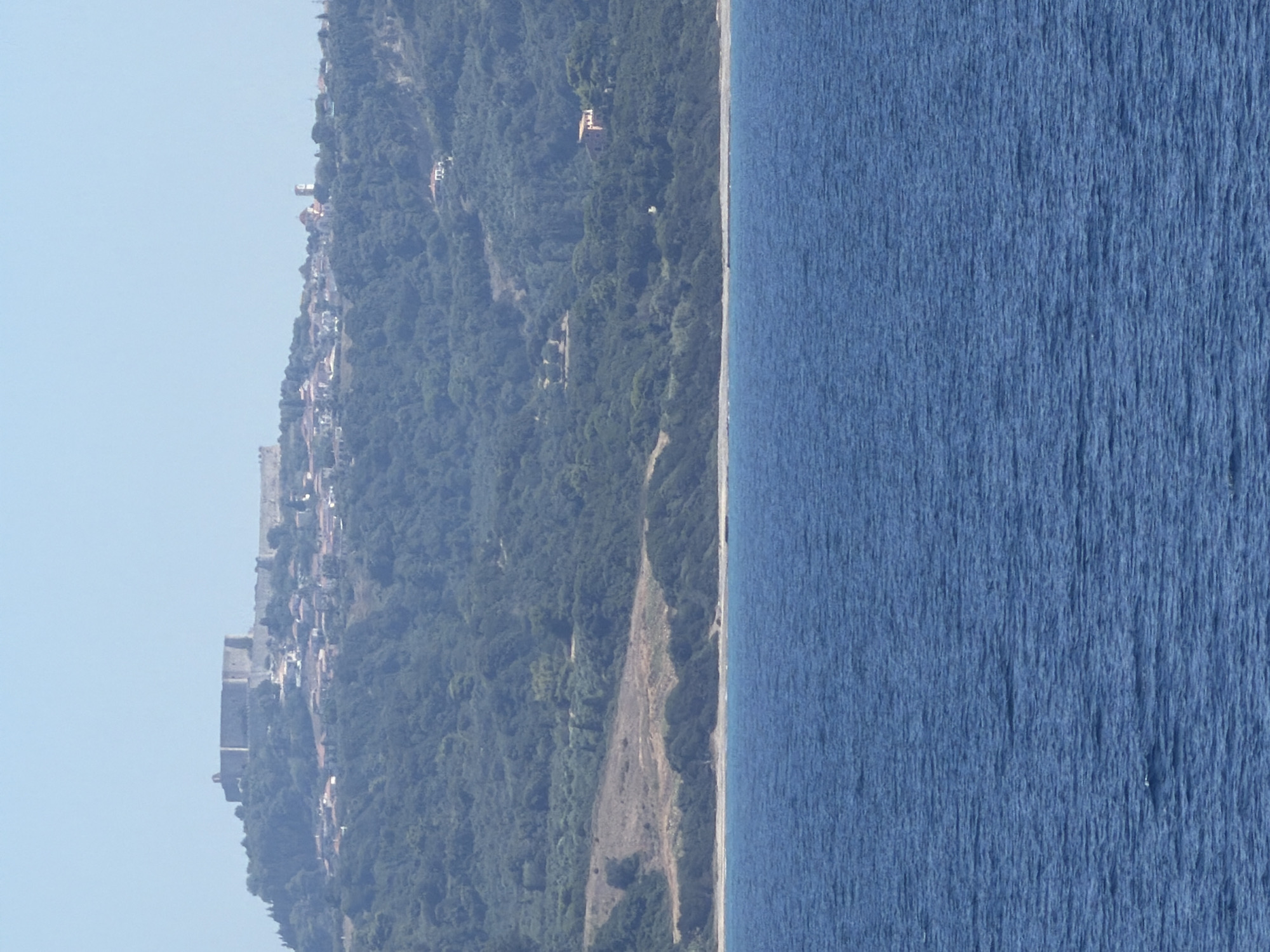 A long shot captures a coastal view from the sea. In the foreground, the deep blue water of the sea is rippling under a clear sky. In the mid-ground, a narrow strip of coastline is visible, featuring dry, sandy terrain and low-lying brush. Beyond the coastline, a densely forested hillside rises. Perched on the crest of the hill, a sprawling ancient fortification dominates the landscape. The stone walls of the fortress are visible, stretching across the hilltop. Below and around the fortress, a village with numerous red-tiled roofs is nestled into the hillside. A prominent church steeple rises above the village. Further down the hillside, a few isolated houses are scattered amongst the trees. The scene is bathed in bright daylight.