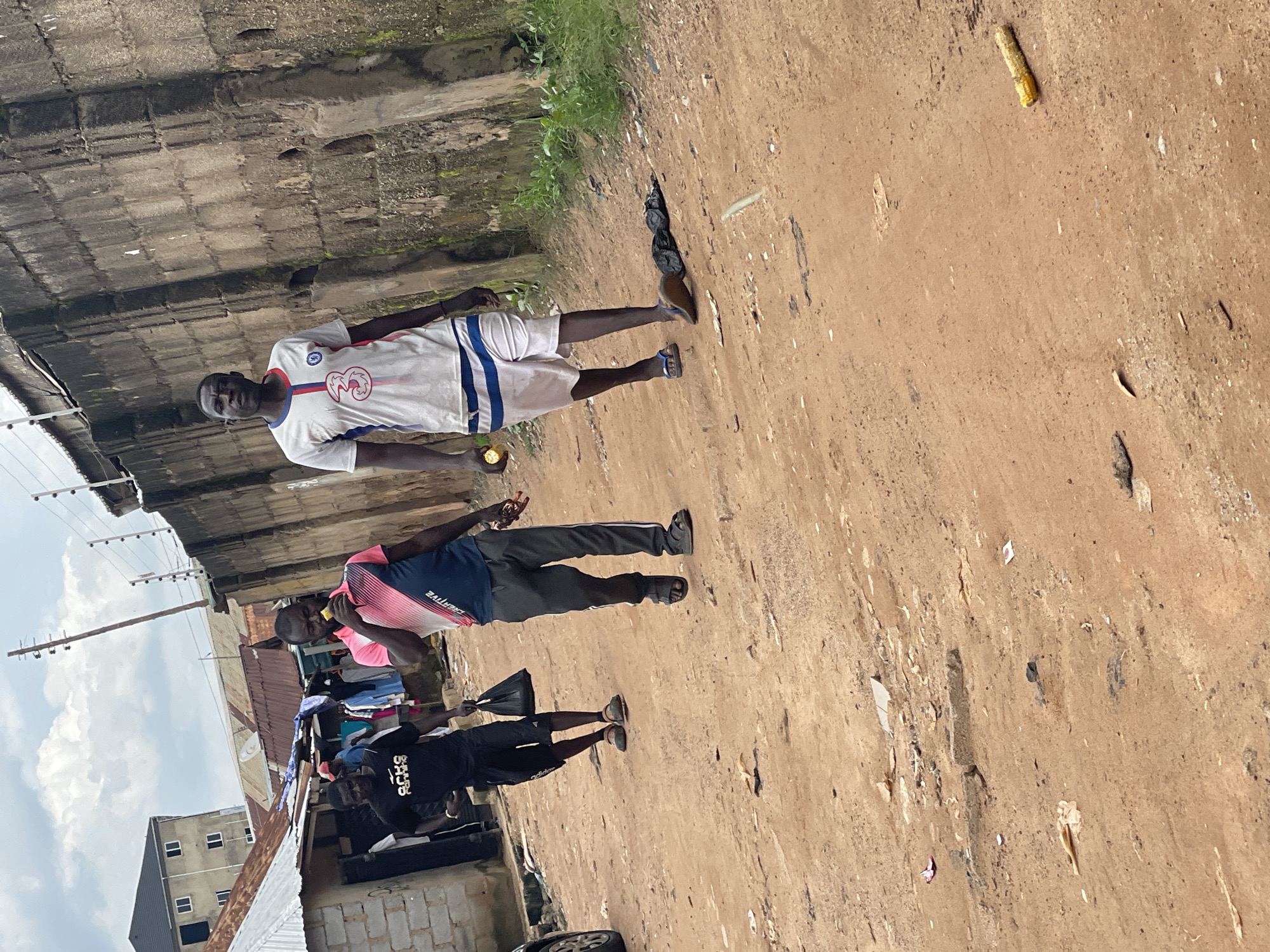 Three men are walking on a dirt road in what appears to be a residential area, possibly in Abuja, Nigeria. The man in the foreground on the right is wearing a white t-shirt with red and blue trim and the number "3" on it, along with white shorts with blue stripes. He is holding a piece of corn to his mouth. To his left, a man in a pink and blue t-shirt is walking and holding a piece of corn, also appearing to eat it. He is wearing dark gray pants. Further to the left, a man in a black t-shirt and black shorts is walking and carrying a black bag. The background shows a high concrete wall with a barbed wire fence on top, and several buildings with corrugated metal roofs. The ground is dusty and uneven, with some scattered debris. The sky is cloudy.