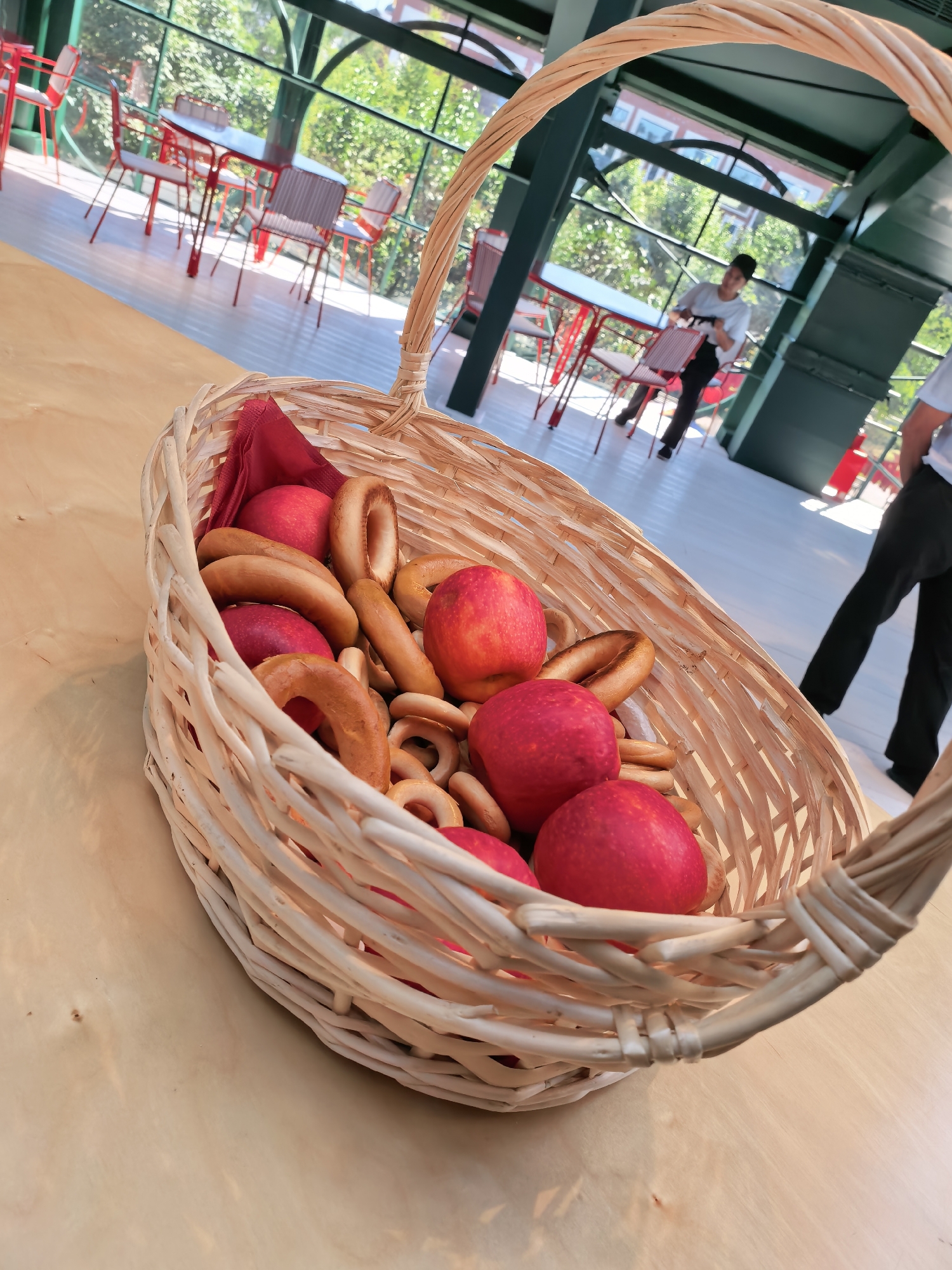 A close-up, high-angle shot shows a wicker basket filled with bagels and red apples on a light-colored wooden table. In the background, a person is visible in a white shirt and dark pants, walking through an outdoor dining area with red tables and chairs under a green awning. The scene appears to be a cafe or restaurant patio.