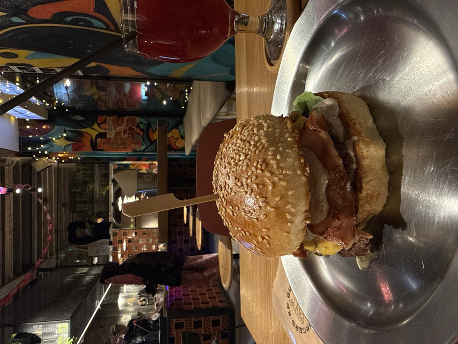 A burger with a sesame seed bun, bacon, cheese, and lettuce rests on a silver plate. A wooden skewer secures the burger's components. A glass of red liquid, possibly wine or juice, stands behind the plate. The background reveals a lively indoor space with string lights, colorful wall art, and other restaurant patrons visible. The scene conveys a casual dining atmosphere.