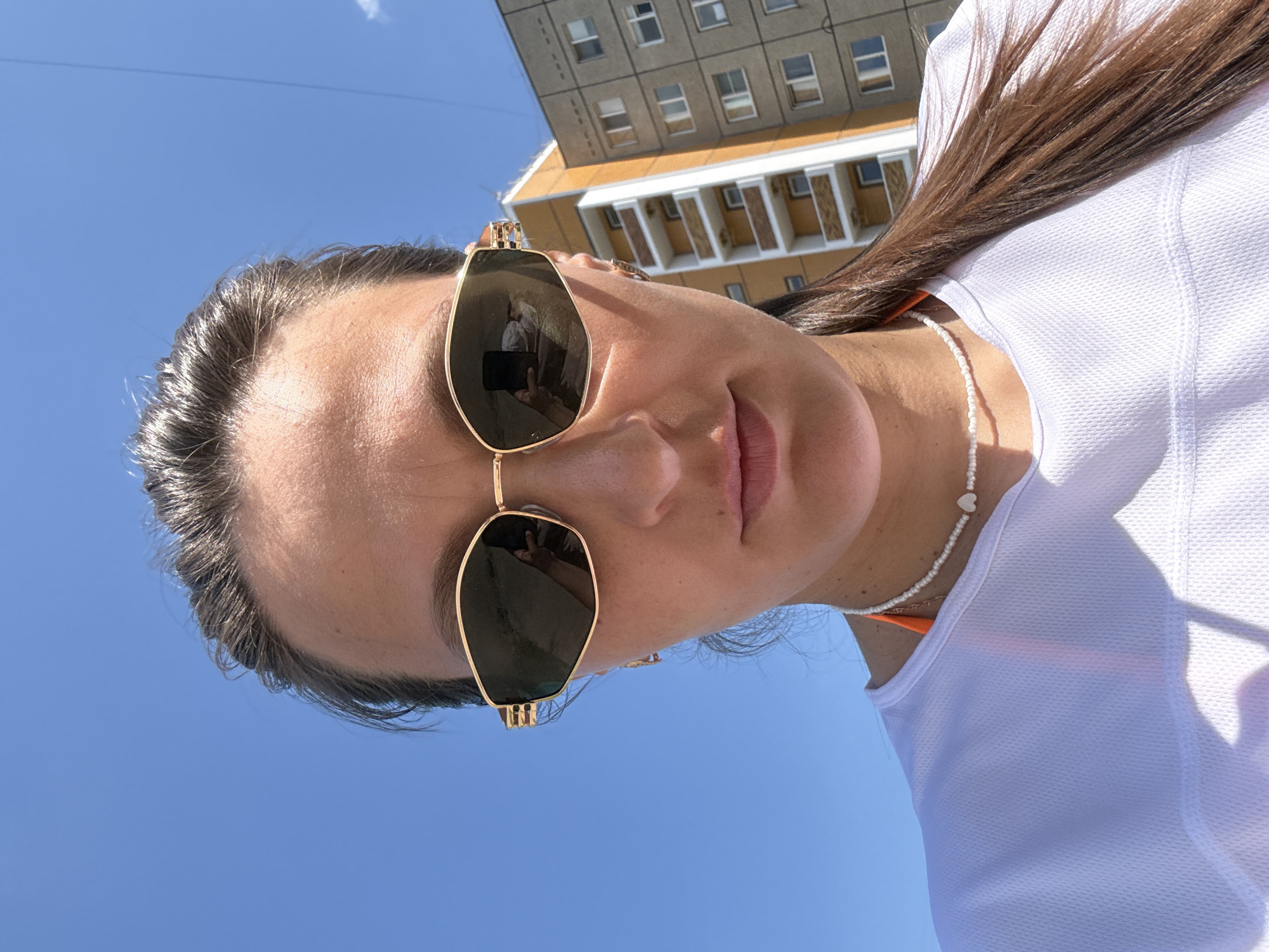A woman with dark hair pulled back and wearing gold-rimmed sunglasses is taking a selfie in front of a grey and orange apartment building under a clear blue sky. She is wearing a white t-shirt and an orange bikini top, with a white beaded necklace with a heart pendant. The sunglasses reflect a cell phone being held by the woman to take the photo. The building appears to be in the city of Chaikovsky, Russia.