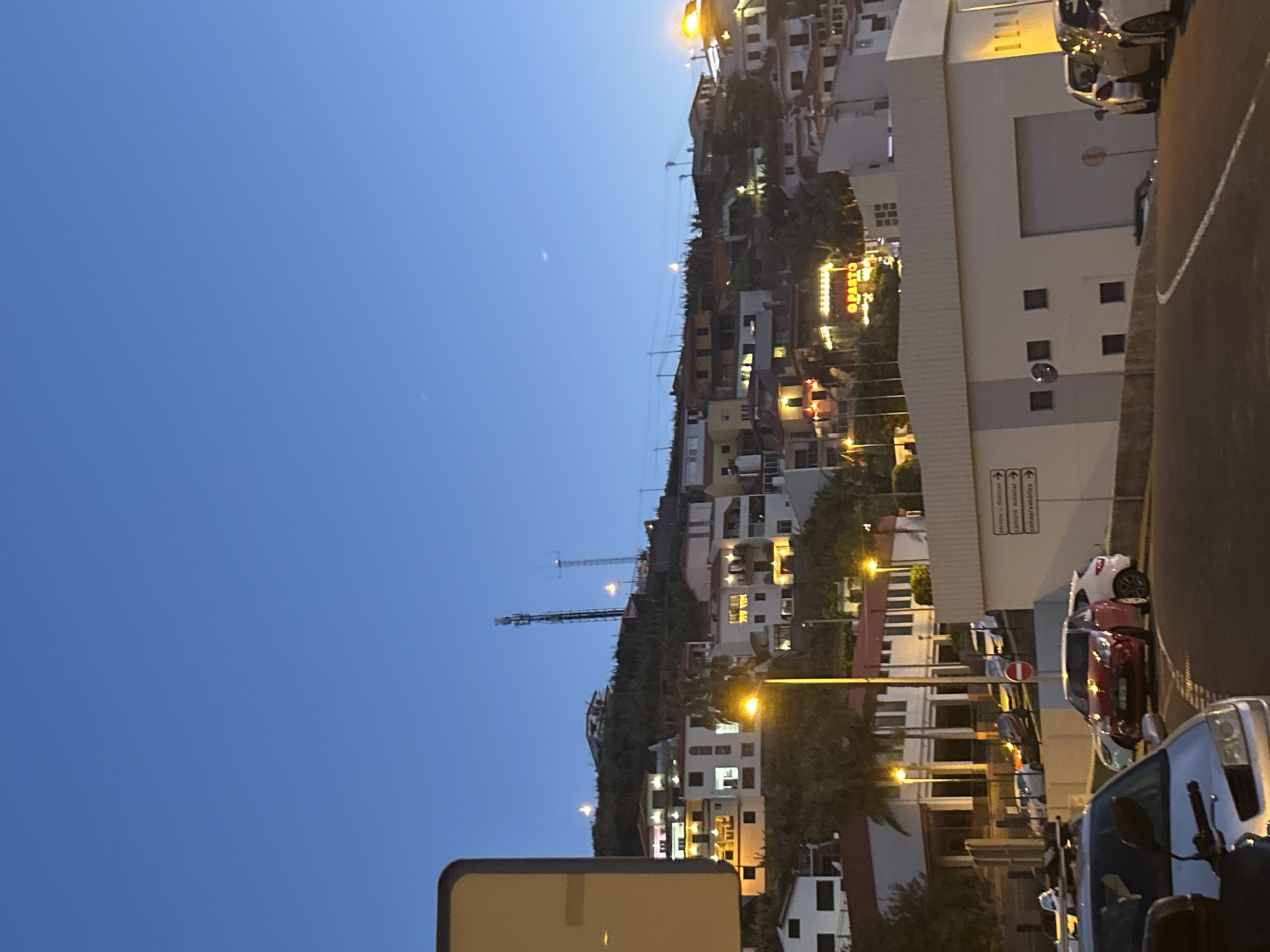 The image depicts a nighttime view in Câmara de Lobos, Portugal. Residential buildings cascade down a hillside illuminated by streetlights and interior lighting. A telecommunications tower is visible in the distance. In the foreground, a street scene includes parked cars, a building with signage, and streetlights.