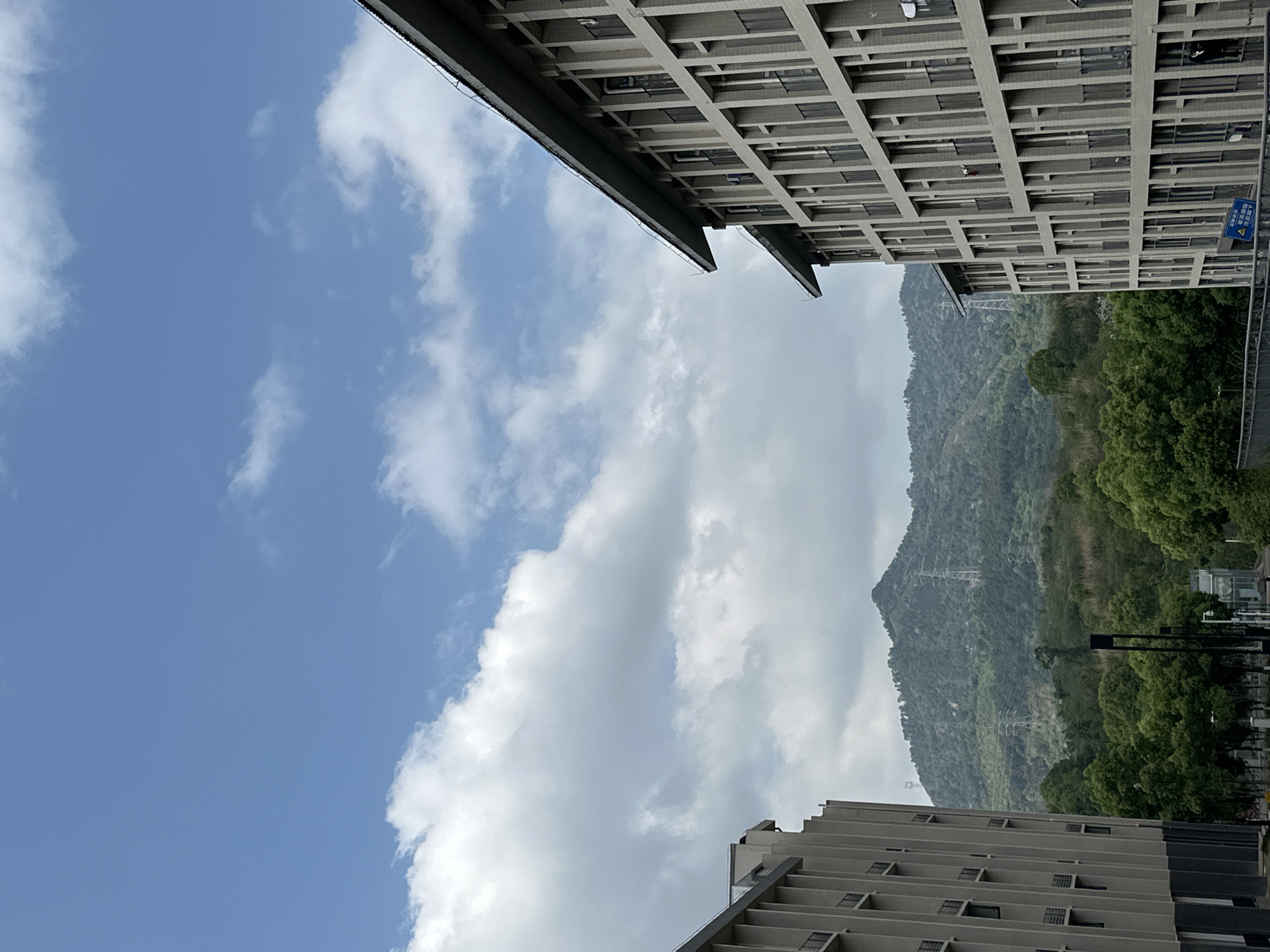 The image presents an upward view framed by the edges of two buildings. The sky is blue with scattered white clouds. A mountain range, covered in dense green vegetation, lies in the background. Electrical transmission lines are visible against the mountain. The location is likely in Jiangjin, China due to the architectural style of the buildings and the presence of the mountain range in the background. There are no people visible.
