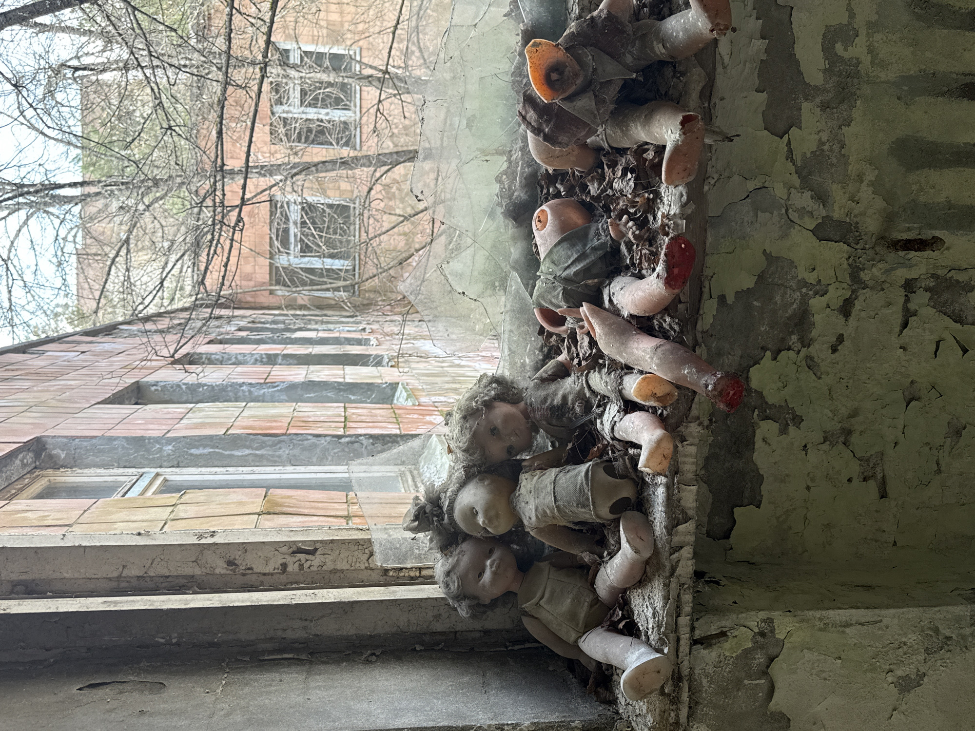 The image depicts a low-angle shot of an abandoned building in what is presumed to be Хутор, Ukraine. The exterior is constructed of decaying brick and concrete, with visible window frames. Close to the viewer, embedded in the wall or a window frame, is a collection of damaged dolls and doll parts, mixed with leaves and debris. The dolls exhibit significant wear and tear, with missing limbs and damaged features. The wall itself is heavily weathered with peeling paint. The angle of the shot allows for a view of the building's facade extending upwards, interrupted by bare tree branches.