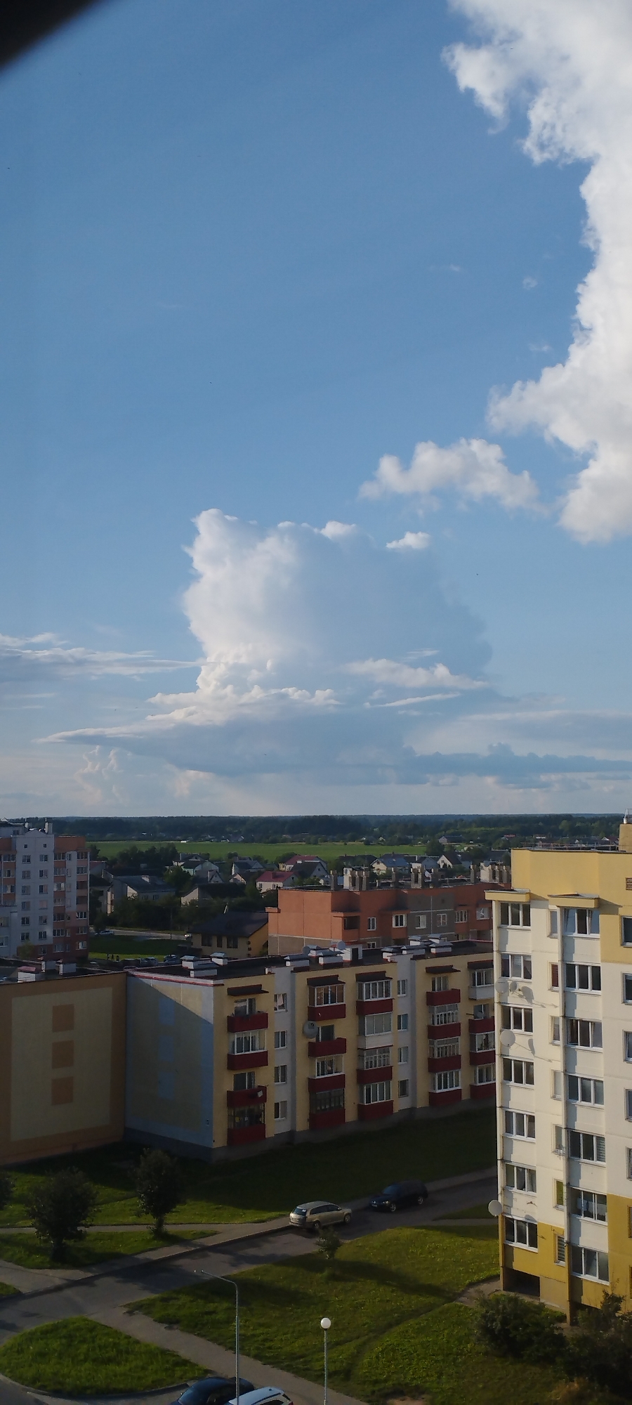 The image shows an aerial view of apartment buildings and a suburban landscape in Skidzieĺ, Belarus. In the foreground, the top portions of several multi-story apartment buildings are visible. These buildings are characterized by their colorful facades, with a prominent yellow and white building on the right, and yellow and red apartment blocks to the left. Balconies with red railings are visible on some of these structures. Beyond the apartment buildings, a strip of green trees separates the urban area from a field of green grass and scattered houses with red roofs, indicative of a suburban or rural setting. The background is dominated by a bright blue sky with scattered white cumulus clouds. The lighting suggests it is daytime, likely late afternoon, with shadows cast by the buildings and trees. There are no visible people in the image. The scene is peaceful and urbanized, with elements of nature present in the surrounding landscape.