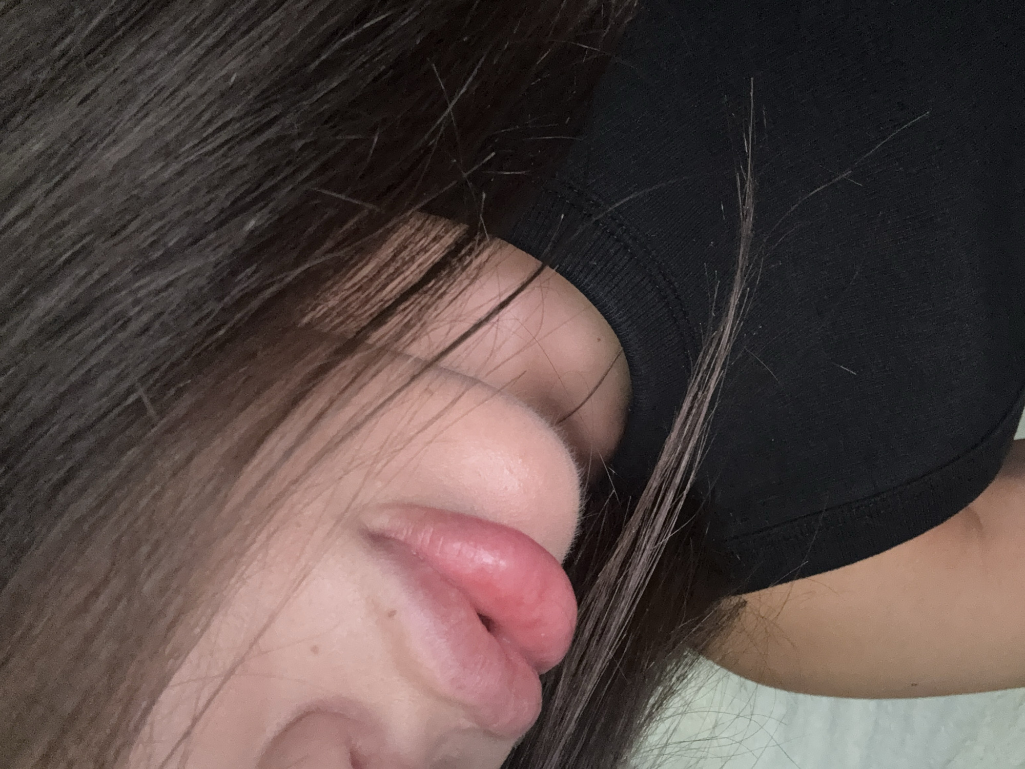 A close-up, head-and-shoulders shot shows a portion of a person's face, including their lips and chin, with dark brown hair framing the left side of the image and resting on their shoulder. The person is wearing a black sleeveless top. The background appears to be a light-colored, textured surface. The lighting is soft, creating a subtle glow on the skin. There are no discernible elements that would place the scene in Basarabeasca, Moldova.