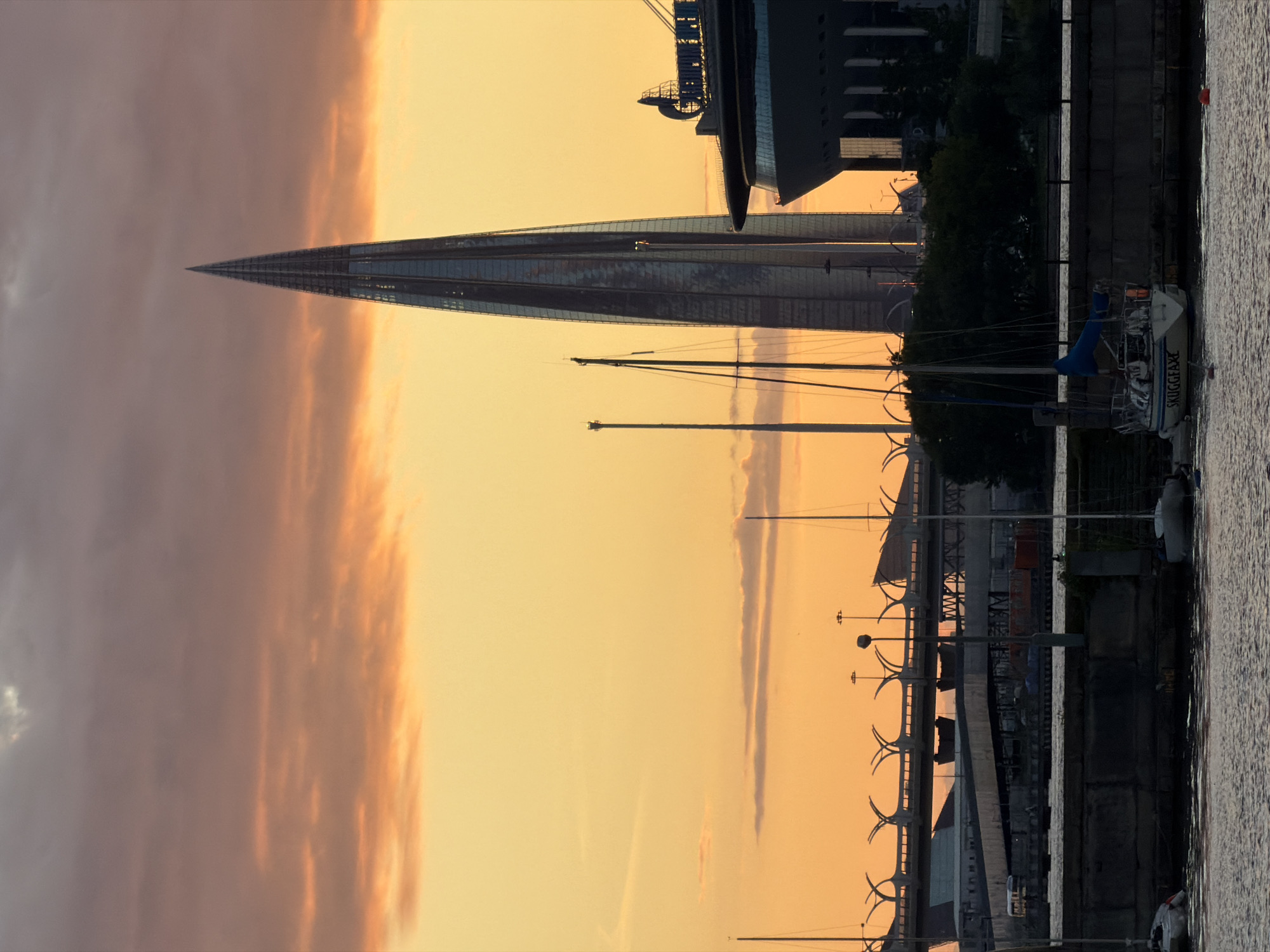 The image depicts a view of Saint Petersburg, Russia, at sunset. Dominating the scene is the Lakhta Center skyscraper, a modern, needle-shaped building with a glass facade, silhouetted against a warm, orange and yellow sky filled with scattered clouds. In the foreground, the Neva River is visible, with several sailboats moored along its bank. One sailboat in the center has "SKUGGEFAXE" written on its stern. A large cruise ship or ferry is docked to the right of the skyscraper. In the mid-ground, a distinctive bridge with curved, arching supports spans across the water. The overall atmosphere is serene, with the fading light casting long shadows and highlighting the architectural elements of the city.