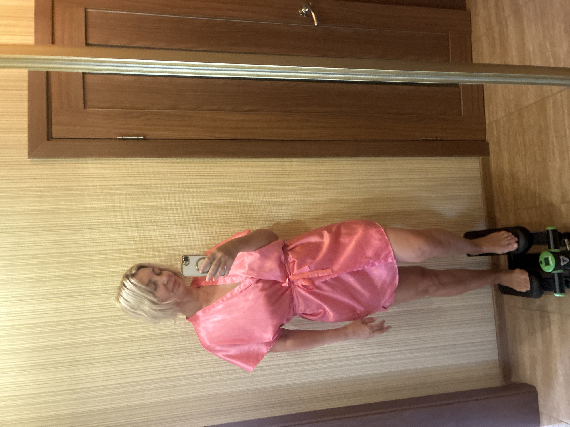 A woman with blonde hair, wearing a pink satin robe, is standing on a stair stepper machine, holding a smartphone in her right hand. She is looking at the phone as if taking a selfie. She is indoors, in front of a beige vertical striped wallpaper. To her right are two wooden doors. The floor is tiled. The image does not provide any specific location information within Sochi, Russia.