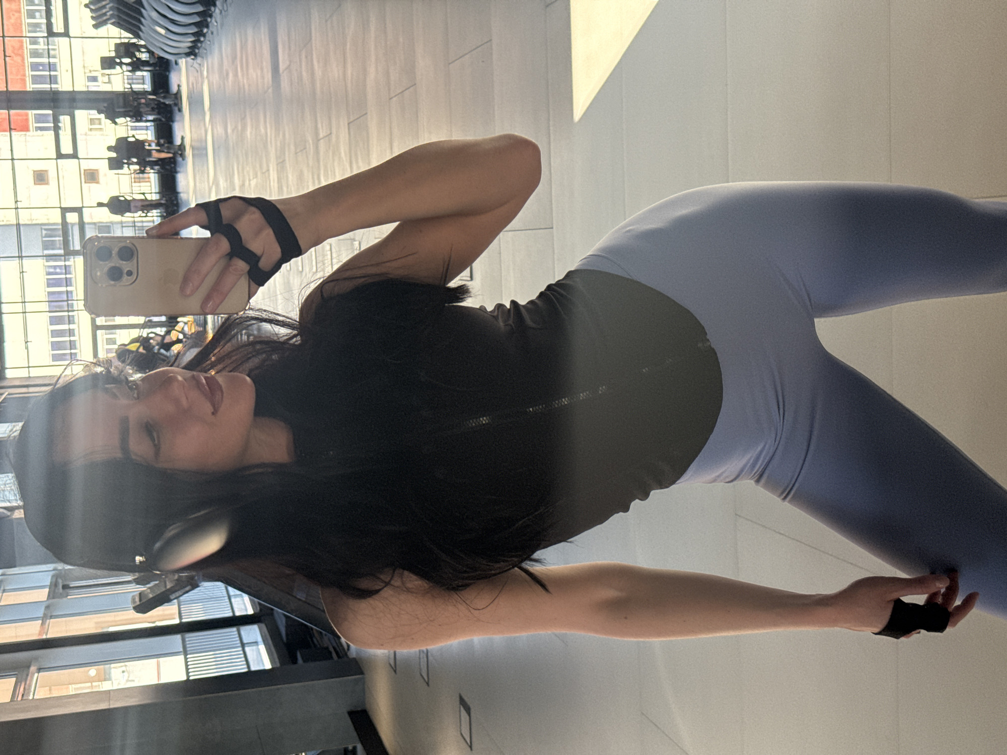 A young woman is captured in a mirror selfie, posing in what appears to be a modern gym or fitness center. She has long, dark hair and is wearing a black athletic top, light blue leggings, and black fingerless gym gloves or hand wraps. Headphones are visible around her neck or ears. She holds a light-colored smartphone (resembling an iPhone) with a multi-lens camera in her right hand, positioned to take the photo. Her expression is a subtle smile or smirk, conveying a relaxed and confident demeanor.
The setting is characterized by a reflective, light-colored floor and large windows that allow bright sunlight to stream in, casting strong reflections and highlights. Through the windows, an urban landscape is visible, featuring multi-story buildings with numerous windows, suggesting a city environment. The reflections on the floor and in the glass also show outlines of gym equipment or seating, reinforcing the gym context. The strong sunlight indicates it is daytime. There is no clearly discernible text visible on any objects or in the background, though the phone appears to be an Apple iPhone. The overall scene suggests a pause during or after a workout in a contemporary fitness facility located in an urban area, likely Krasnoyarsk, Russia, as per the provided context.