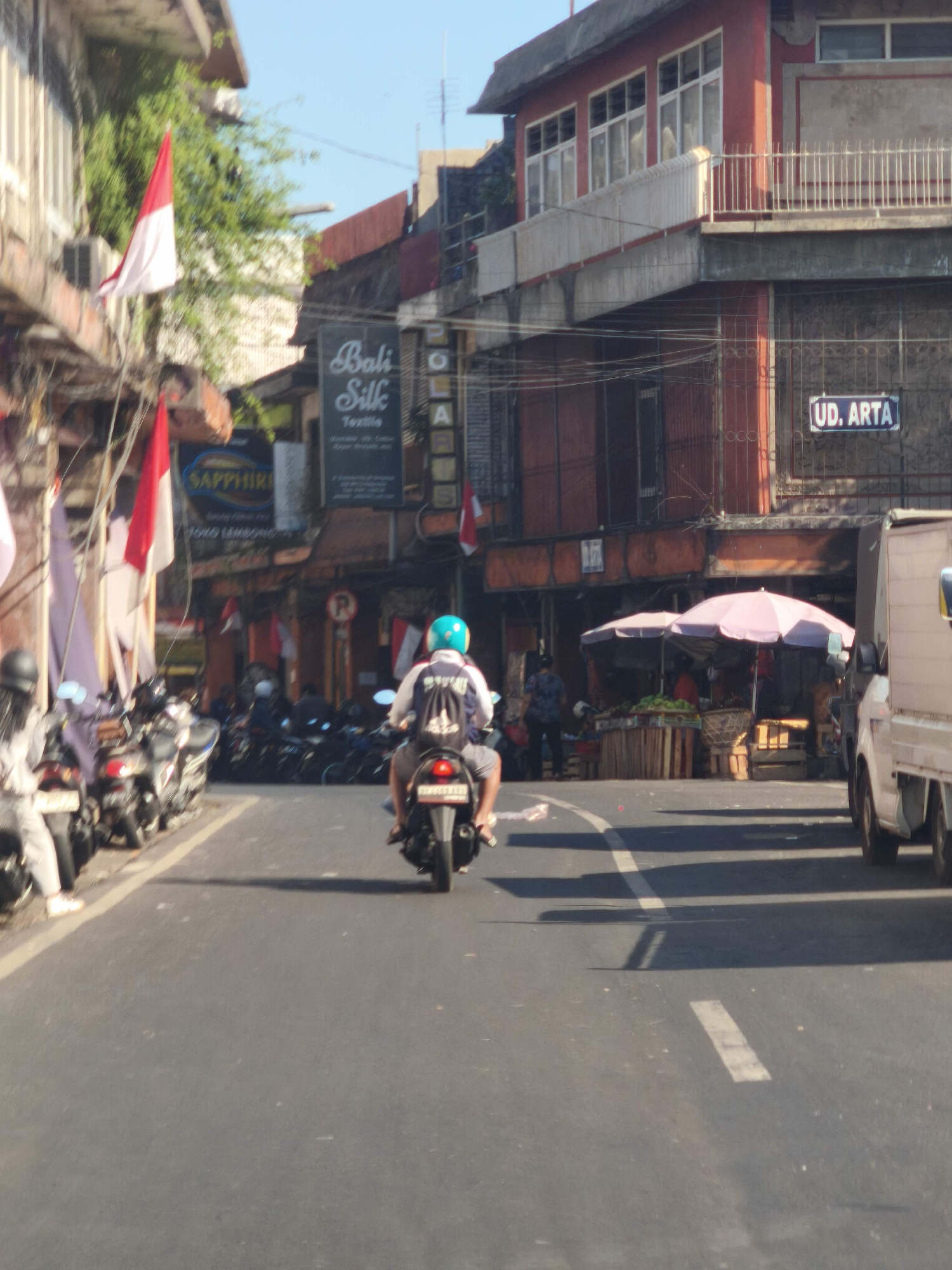 In the city of Denpasar, Indonesia, a street scene unfolds with several motorcycles parked along the left side of the asphalt road. A person wearing a blue helmet and a backpack is riding a black motorcycle in the center of the frame, heading away from the viewer. To the right, a white truck is visible, and beyond that, market stalls with pink and purple umbrellas display produce. Buildings line the street, some adorned with Indonesian flags. A sign for "Bali Silk Textile" is prominent on one of the buildings. Another sign displays "UD. ARTA". The sky is clear and blue.
