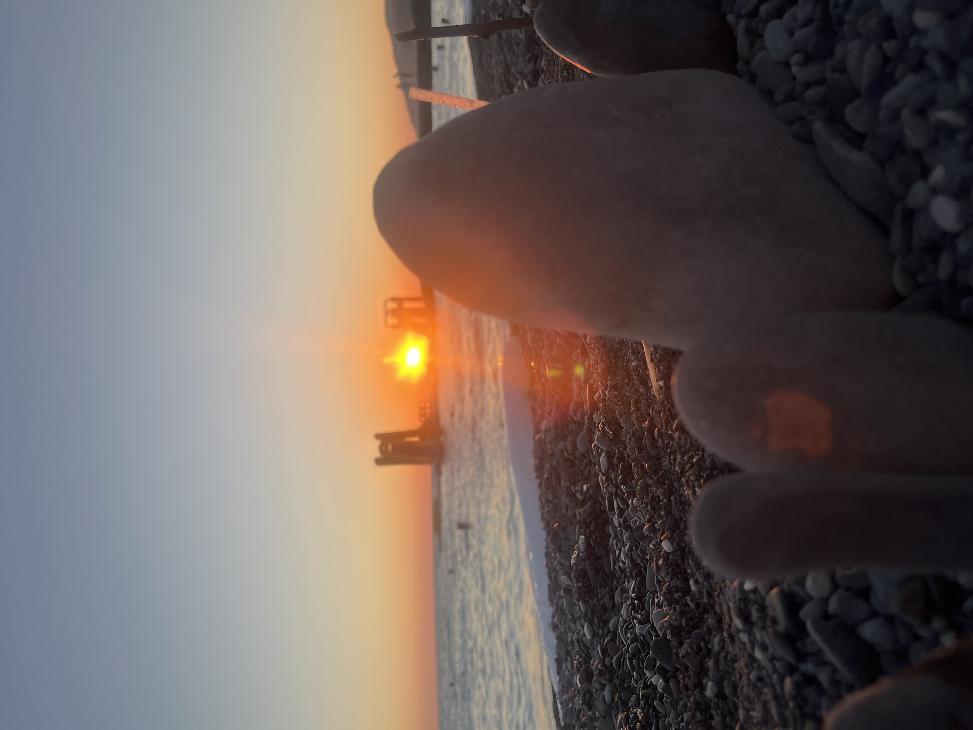 The image depicts a sunset on a pebbled beach in Sochi, Russia. The sun, a bright orb, is low on the horizon, casting a warm orange glow across the sky and water. In the middle ground, a yellow water slide structure is visible in the sea. The foreground is dominated by smooth, grey pebbles, with what appear to be two toes or a pair of feet, also appearing as rounded stones, positioned in the lower left of the frame. Another large, smooth stone is positioned in the center right, and a small stick is visible behind it. The sea is calm, with gentle waves lapping at the shore. The sky transitions from a pale blue at the top to a fiery orange and yellow near the horizon. The overall impression is one of tranquility and a picturesque end to the day.