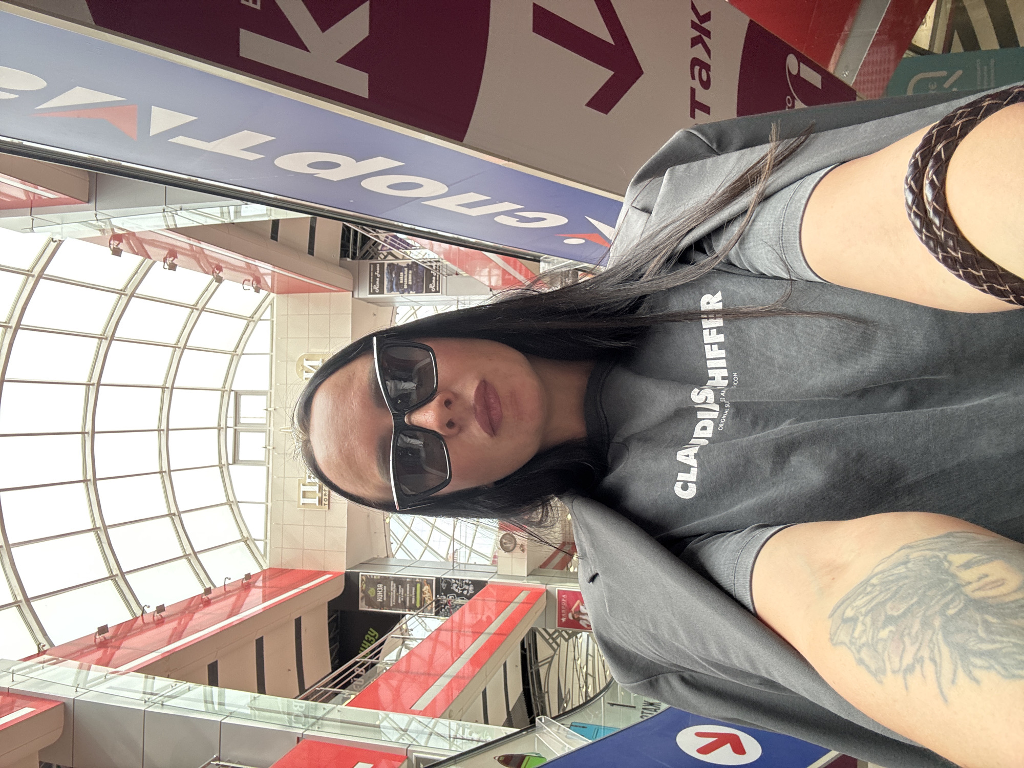 The image shows a woman with dark hair, wearing sunglasses and a gray t-shirt with the text "CLAIDIS HIFFER ORIGINAL SU ICON". She has a tattoo on her arm and a braided leather bracelet. The background suggests she is in a shopping center or airport. Signs in Russian are visible, including "спорTAL" (likely related to sports) and "таж" (likely related to luggage). The location is Izhevsk, Russia, based on signage in the image. The architecture includes a curved glass ceiling and multi-level interior, with escalators and walkways.
