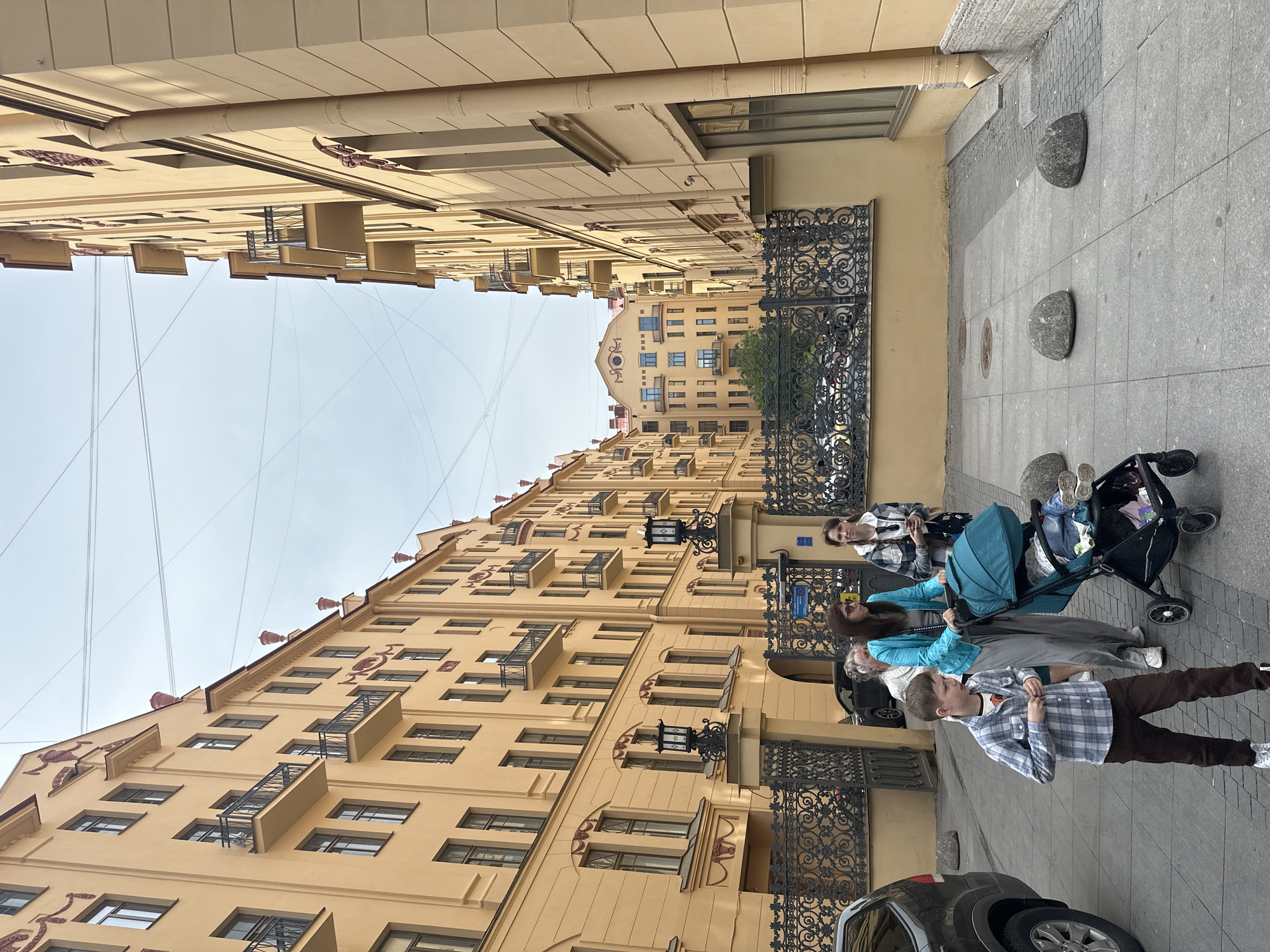 An outdoor urban scene is depicted, likely within Saint Petersburg, Russia, identifiable by the architectural style of the buildings. The tall structures on either side of the street are painted in a light yellow hue and feature symmetrical window placement along with decorative wrought iron balconies. Electrical lines stretch across the sky overhead.
On the right side of the frame, a sidewalk is visible, and it features irregularly placed, smooth grey stones. A dark-colored vehicle is parked on the street, with only its front portion appearing in the bottom right corner of the frame. A family is present on the sidewalk; the composition includes a child in a checkered shirt and brown pants, a woman in a blue jacket, and another woman standing behind a stroller. The stroller has a blue cover. The subjects appear to be casually strolling along the sidewalk.