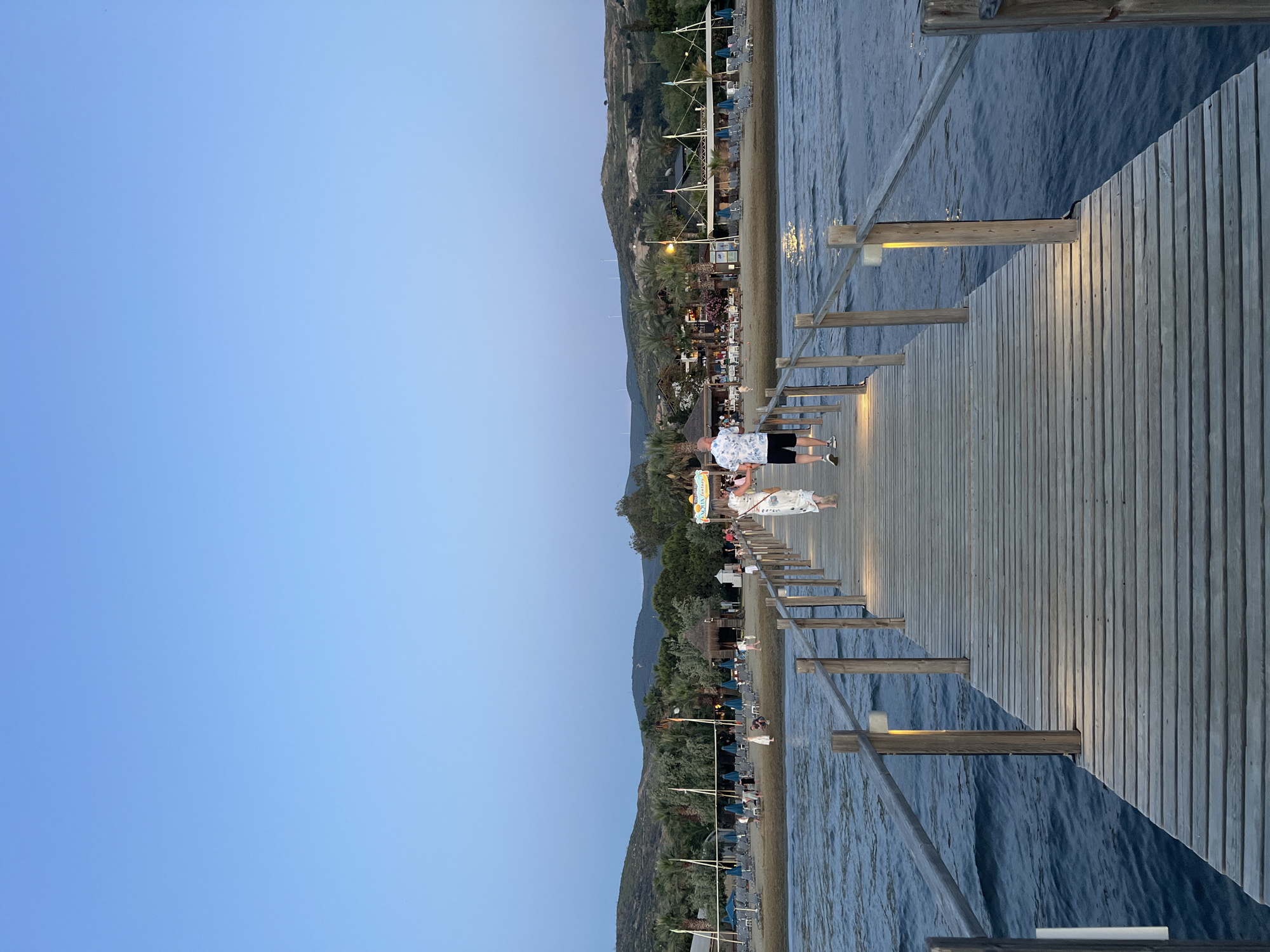 An outdoor image captures a serene coastal scene at dusk or early evening. A wooden pier extends from the left side of the frame into dark blue, slightly rippled water on the right. Small, warm-toned rectangular lights are affixed to vertical posts along the pier, casting reflections onto the water.
On the pier, a man in a patterned short-sleeved shirt and dark shorts walks away from the camera, followed by a woman in a white dress. Further down the pier, additional figures are visible. The left side of the image features a sandy beach lined with sun loungers and umbrellas. Behind the beach, several buildings, possibly restaurants or beach clubs, are illuminated with artificial lights. Palm trees and other greenery are interspersed among the structures. Rising behind the beach and structures are large, green, sparsely vegetated hills. The sky above is a clear, light blue, transitioning from a brighter hue near the horizon to a deeper tone higher up.
The scene depicts a leisurely evening atmosphere, with people strolling on the pier and relaxing on the beach. A sign near the beach area is partially visible with the text "AQUA FANTASY." The overall impression is one of a tranquil resort setting in Selçuk, Turkey, as natural light fades and artificial lights begin to dominate.
