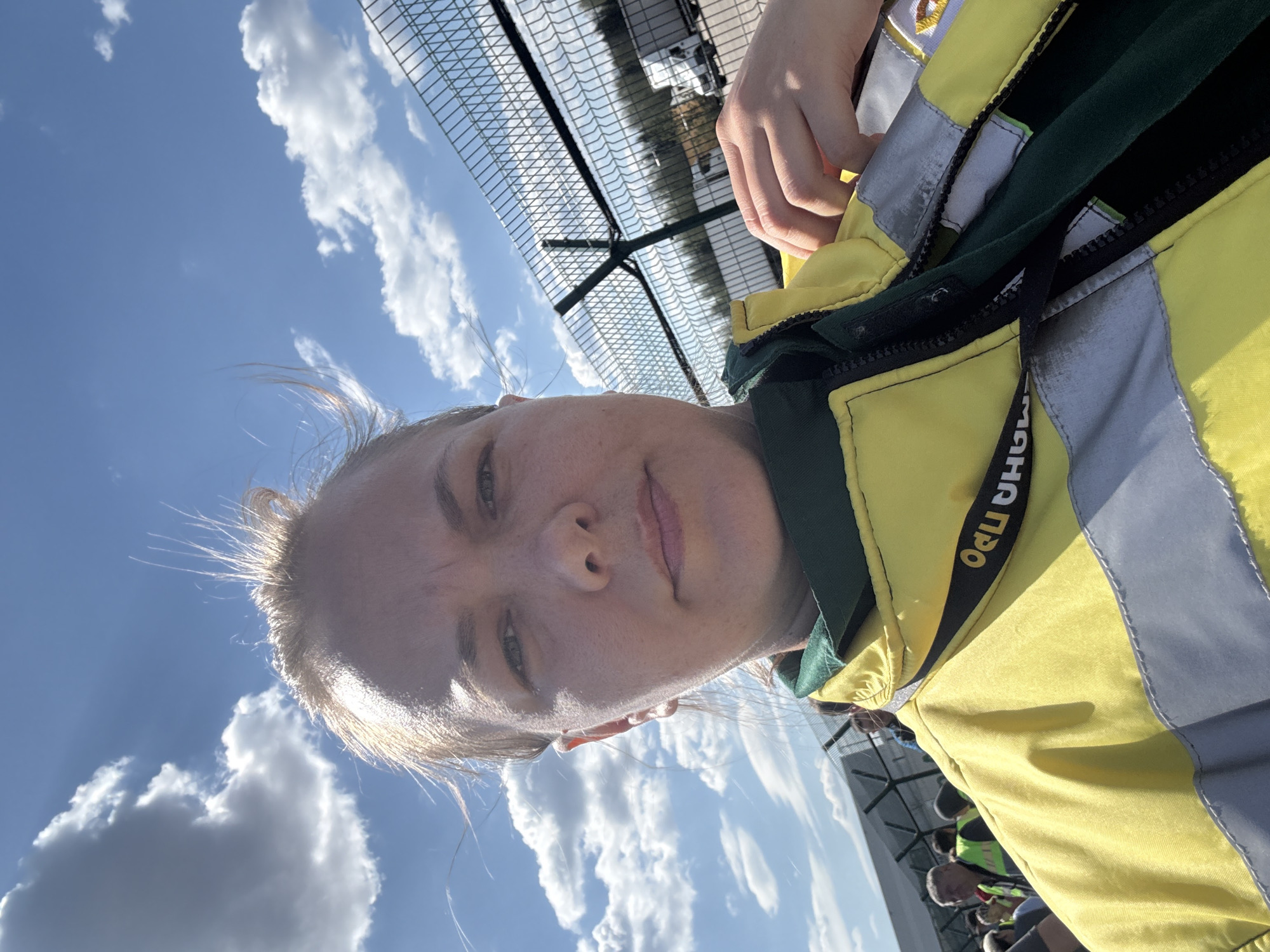 A selfie shows a person with fair skin and blond hair, wearing a yellow and green high-visibility jacket with reflective grey stripes. The name "PPO MAHA" is printed on the jacket. The background includes a blue sky with fluffy white clouds and a chain-link fence. Other individuals can be seen in the background. The location is Lupanovo, Russia.