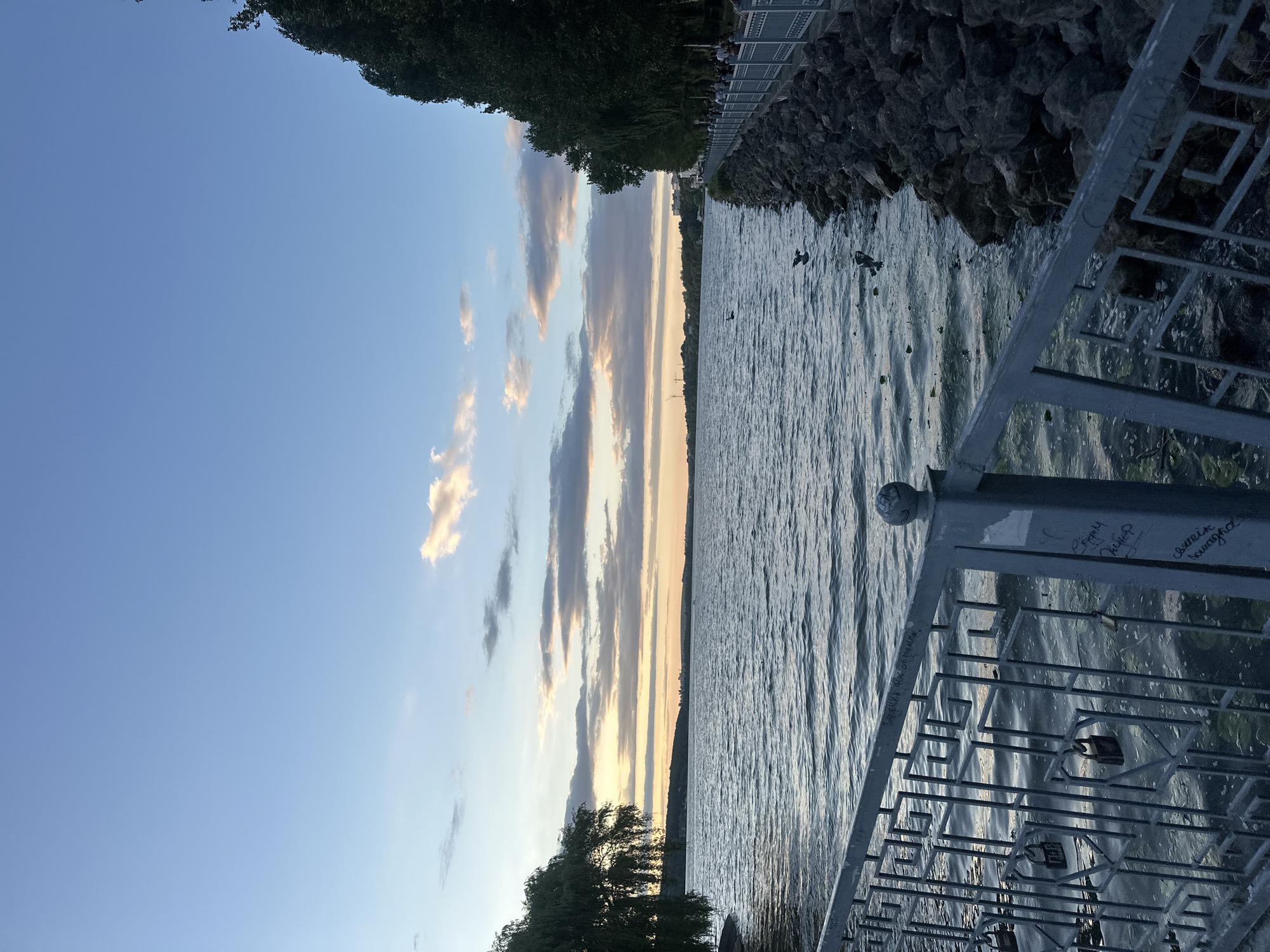 The image depicts a serene waterscape in Ternopil, Ukraine, likely at sunset. The foreground features a metal railing adorned with several padlocks, a common symbol of love. Beyond the railing lies a calm body of water, the Ternopil Pond, with gentle ripples reflecting the soft light of the sky. Along the right bank, a stone embankment is lined with trees, creating a natural border. A promenade or walkway runs parallel to the embankment, where a few individuals can be seen strolling. In the distance, the opposite shore of the pond is visible, characterized by a tree-lined horizon with hints of urban structures. The sky is a canvas of soft blues and pastel hues, with scattered clouds catching the warm glow of the setting sun, creating a tranquil and picturesque atmosphere.
