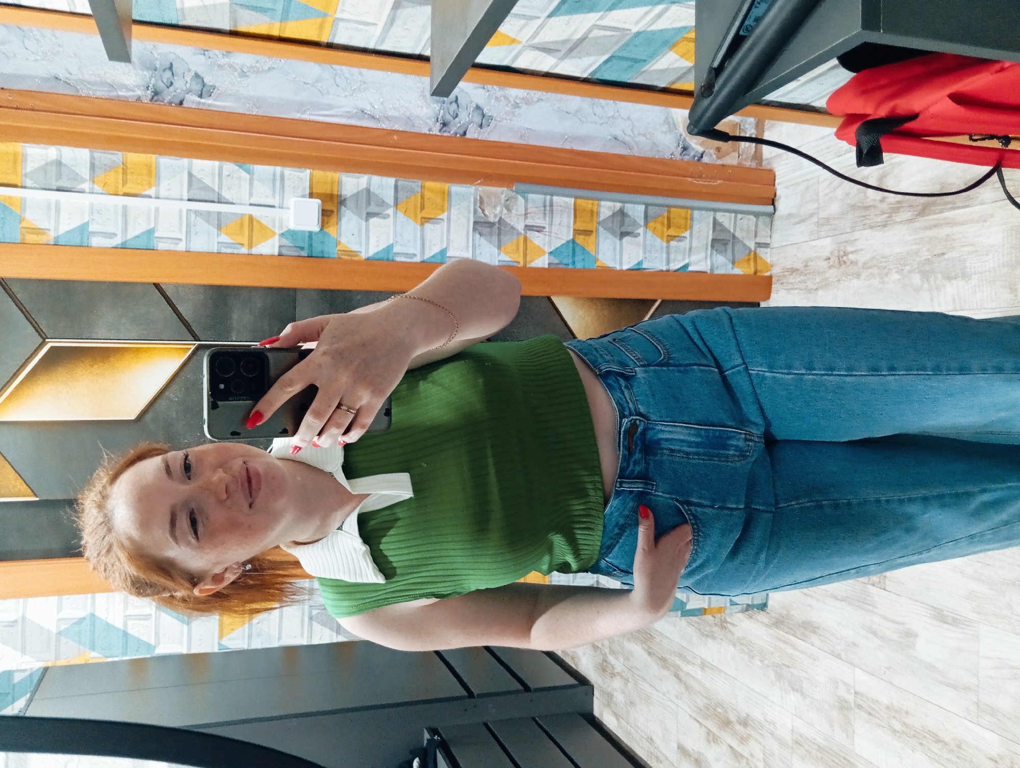 A woman with red hair and freckles is taking a selfie in front of a mirror. She is wearing a green, ribbed shirt with a white collar and blue jeans. Her left hand is in her pocket. She is holding a phone in her right hand with red nail polish. The background shows a wall with geometric patterns in shades of blue, yellow, white, and gray, separated by wooden strips. Part of a dark gray object, potentially a piece of furniture, is visible in the foreground. Another dark gray object with a red item on it is also in the background. The floor is light-colored with a wood-grain pattern. This image was likely taken indoors, in an establishment or residence, based on the architectural elements. Given the user's prompt, it is assumed to be located in Yurgamysh, Russia, but this cannot be confirmed from the visual information alone.