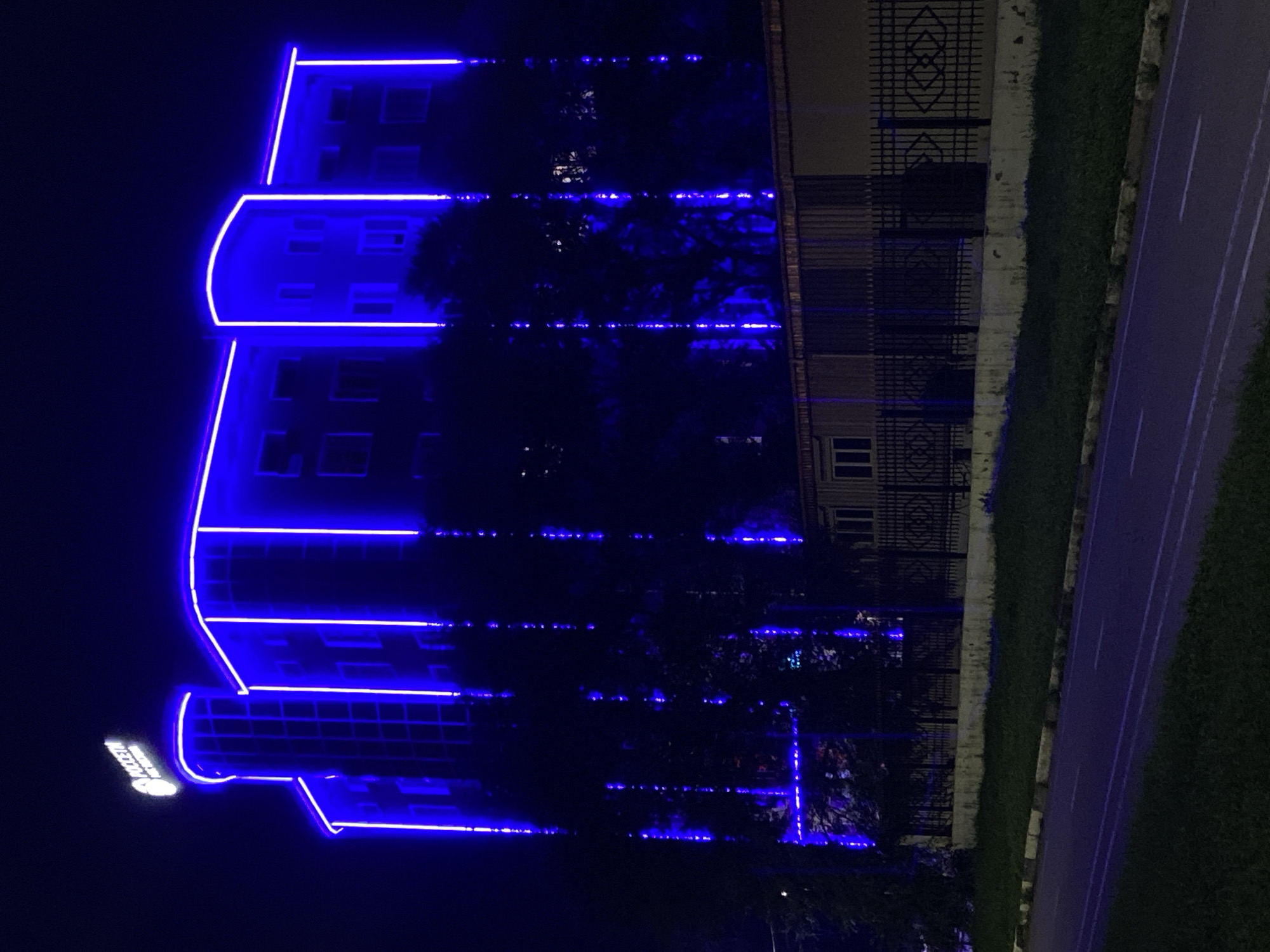 The image depicts a modern building illuminated by bright blue neon lights against a dark night sky. The building has multiple stories and a distinctive curved section on the left side. Neon lights outline the edges of the building, highlighting its architectural features. A sign with text in white and a logo is visible at the top left of the building. In front of the building, there is a black metal fence with an ornamental pattern. Beyond the fence, a long, low building with windows is visible, possibly a storage or utility structure. The foreground shows a paved road with white markings, bordered by a strip of green grass. The scene is located in Blagoveshchensk, Russia, and is likely a commercial or residential complex at night.