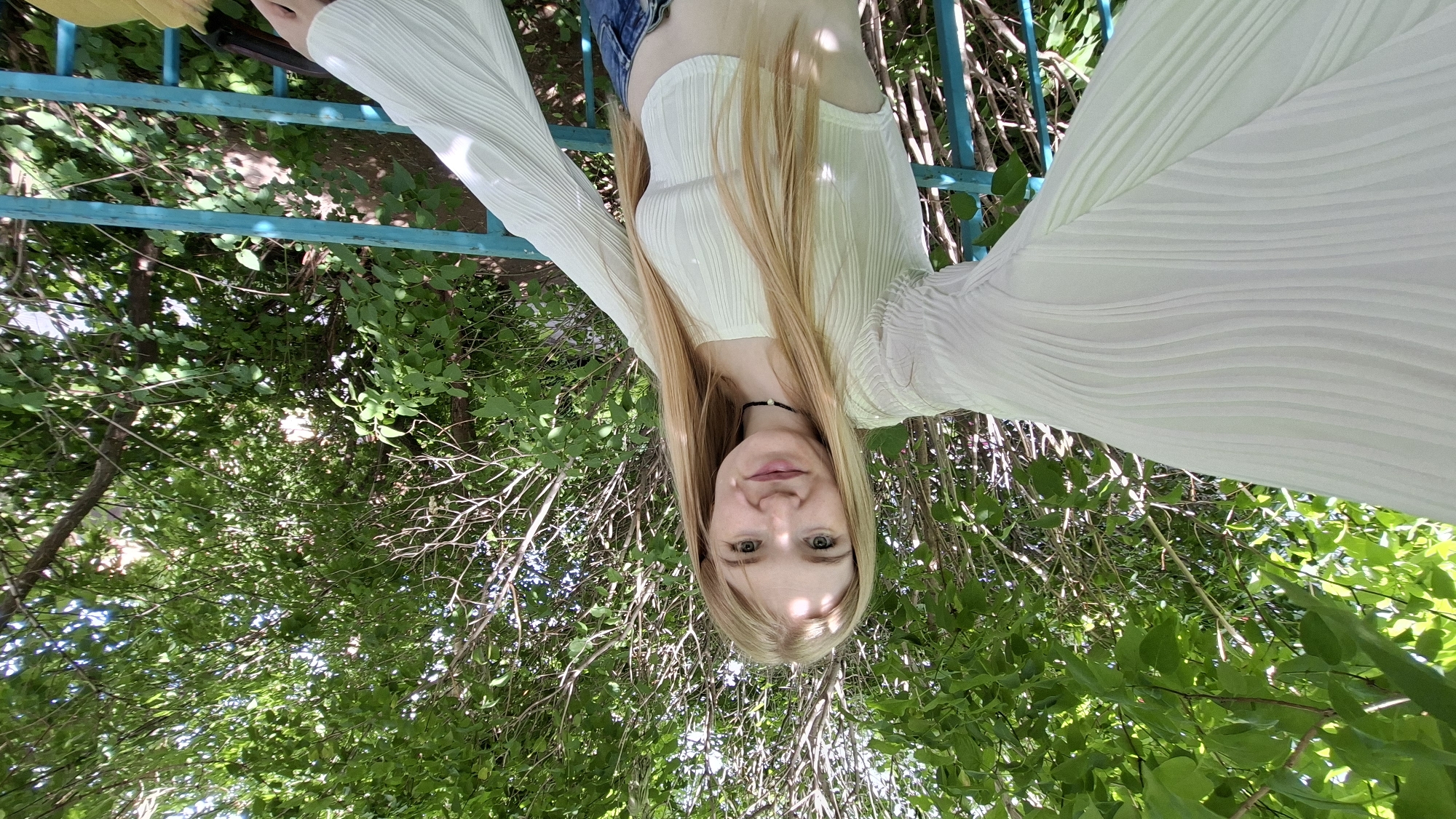 In Chaikovsky, Russia, a young woman with blonde hair and light-colored eyes is captured in a selfie outdoors. She is wearing a light-colored, long-sleeved, ribbed crop top and blue jeans. A black choker necklace adorns her neck. The background features lush green foliage from trees and bushes, suggesting a park or a residential area with greenery. A blue railing is visible in the foreground. The lighting indicates it is daytime, with sunlight filtering through the leaves, creating dappled shadows.