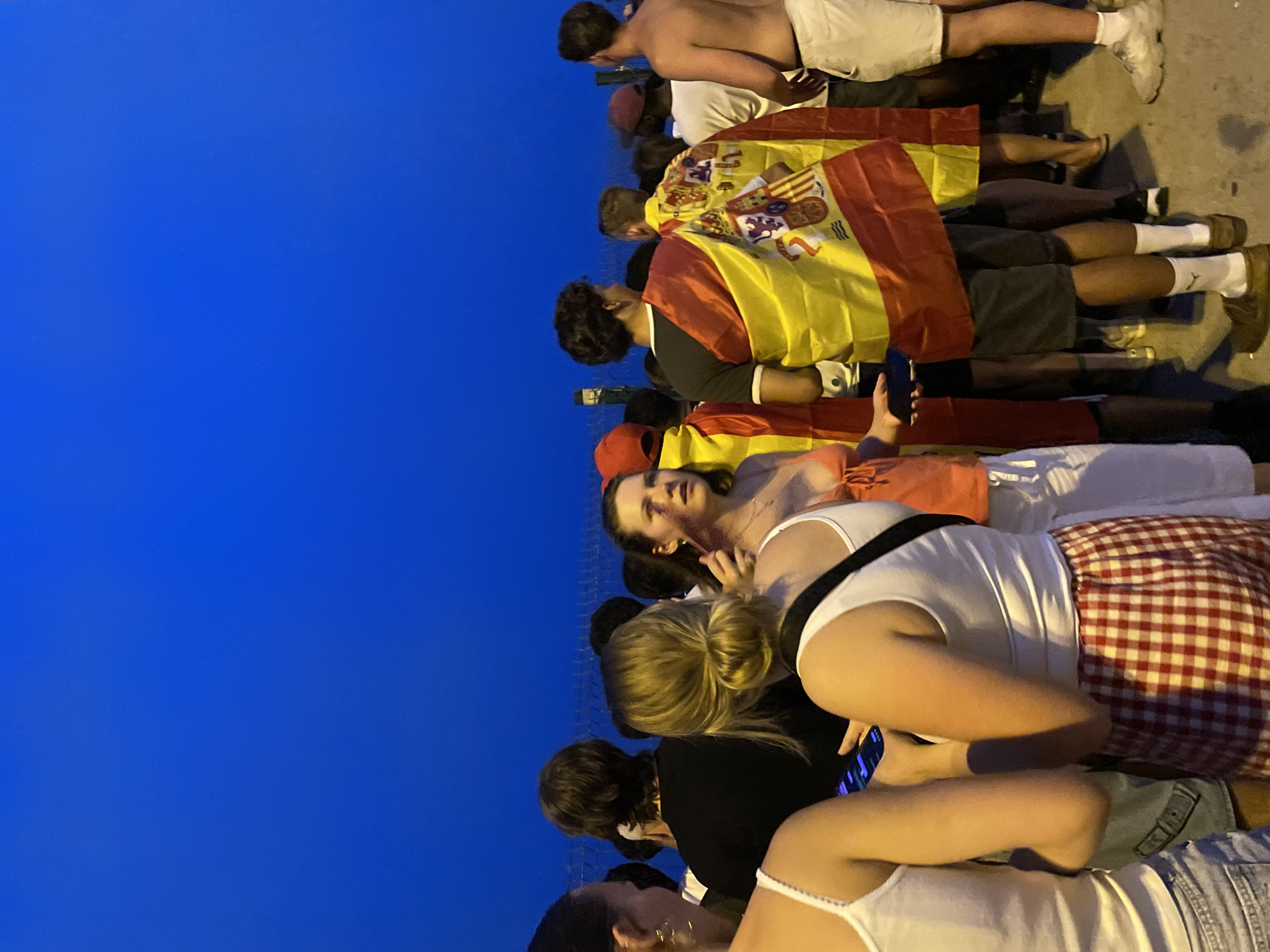 A medium shot captures a crowd of people standing outdoors at dusk against a deep blue sky. The scene appears to be a public gathering or event, possibly a celebration. Several individuals are draped in Spanish flags, the prominent red and yellow colors creating a festive atmosphere. One person in the foreground on the right is shirtless, wearing white shorts and athletic shoes, suggesting an informal setting. Another person in the center-left is wearing a white tank top and red and white checkered shorts, holding a dark-colored object, likely a smartphone, in their hand. The overall impression is one of a communal experience, with people united by national pride or shared excitement. The location is identified as Barcelona, Spain.