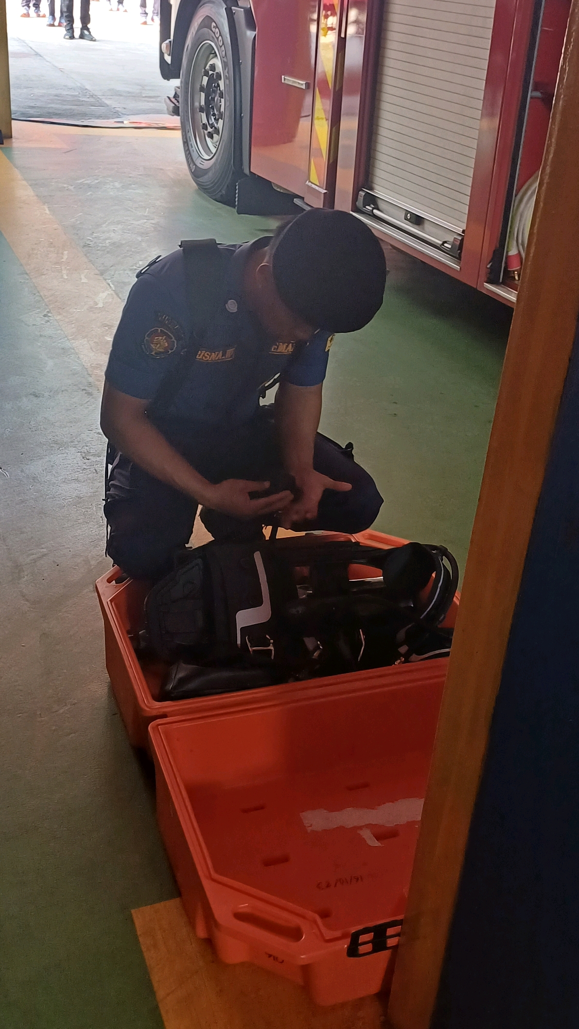 A man wearing a dark blue uniform and a cap, possibly a firefighter, is kneeling next to an open, bright orange plastic case. The case contains a black bag with white accents, which appears to be a piece of equipment, possibly for rescue operations. The man is looking down at his hands, which are positioned over or inside the black bag, suggesting he is handling or inspecting something within it.
In the background, a large red vehicle, likely a fire truck, is visible with a prominent tire and part of its rolling shutter door. The floor is a mix of green and yellow-striped linoleum. The scene appears to be indoors, possibly a fire station or garage, located in South Jakarta, Indonesia. Other individuals in uniforms are partially visible in the far background, standing near the vehicle.