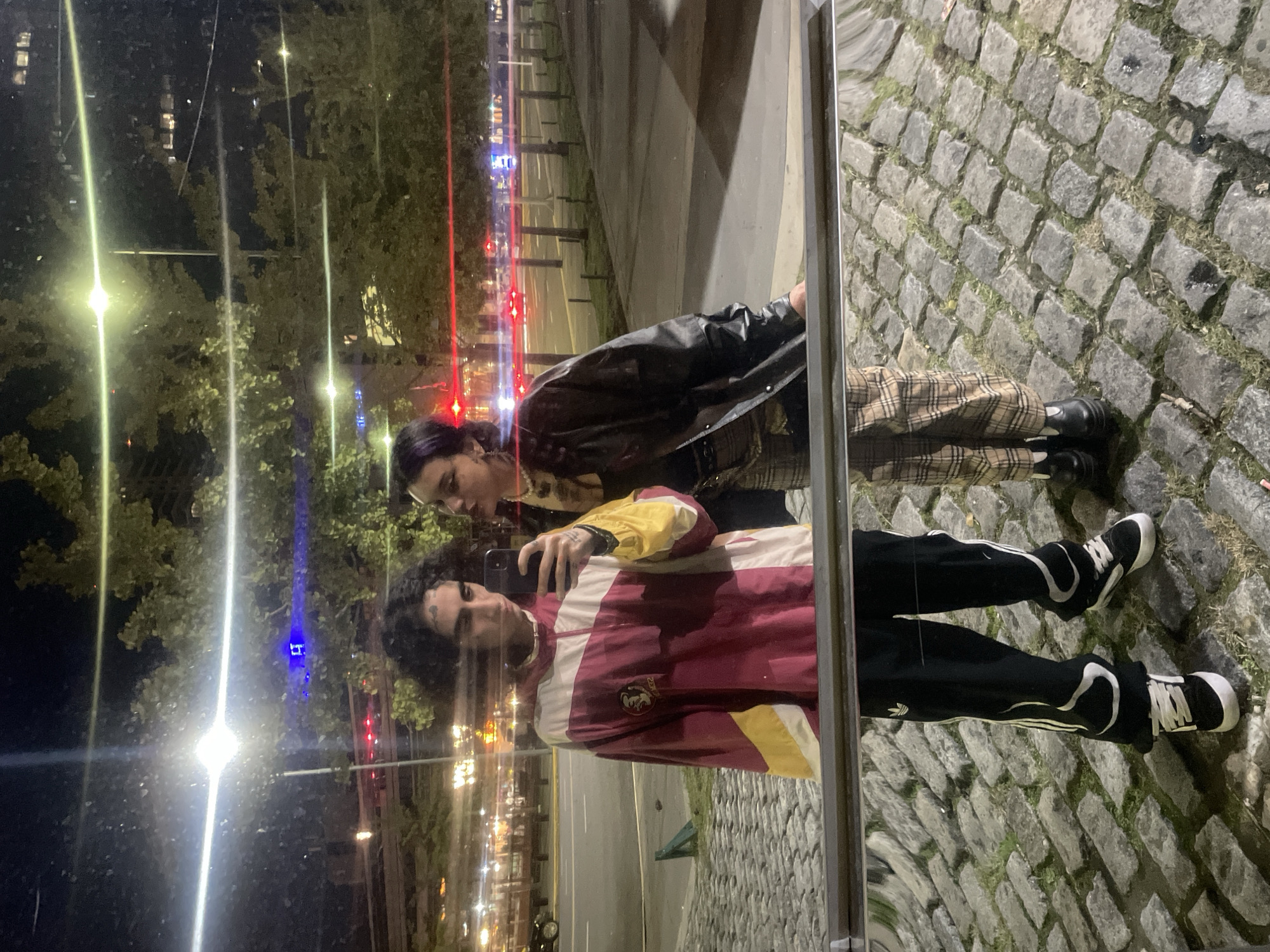 A mirrored image shows two individuals standing next to each other on a cobblestone sidewalk. The person in the foreground, presumably taking the photo, is wearing a red, white, and yellow striped jacket with black track pants and black and white shoes. The second person is wearing a black leather jacket and plaid pants, also with black shoes.
The reflection reveals an urban environment with streetlights, trees, and buildings in the background. The presence of lights and architecture is consistent with a city setting.
Given the cobblestone sidewalk, this appears to be in a historical district of Buenos Aires, Argentina.