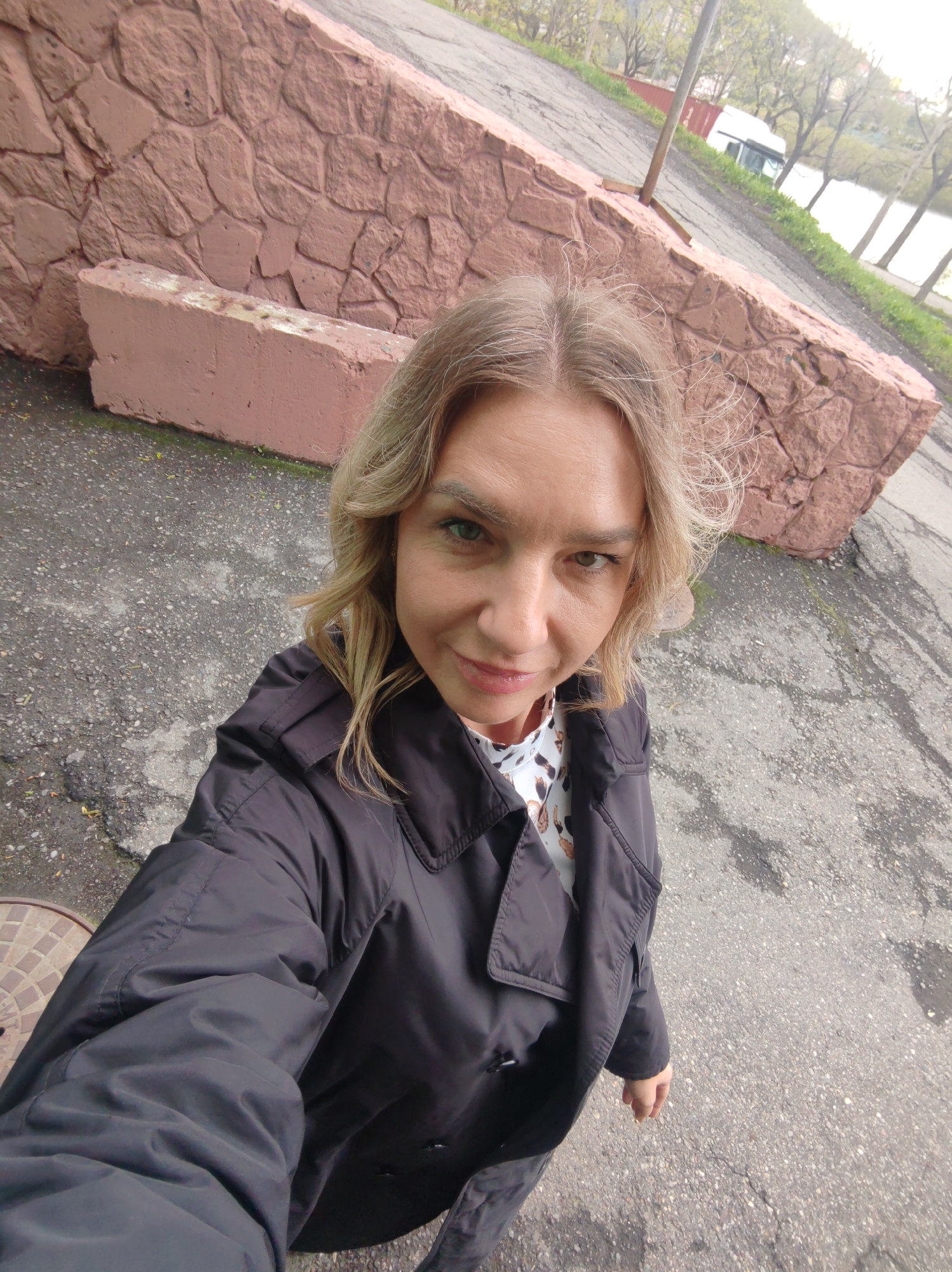 A woman is taking a selfie outdoors. She has blonde hair and is wearing a black trench coat over a printed blouse. Behind her is a pink wall made of large stones. The ground is asphalt. In the distance, trees, a body of water, and a white vehicle can be seen. The location appears to be in Petropavlovsk-Kamchatsky, Russia.
