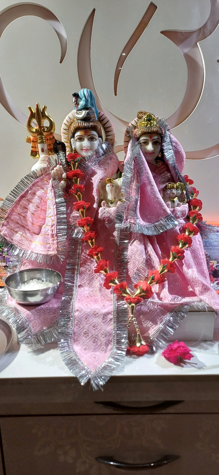 The image displays a religious altar in a home or a small shrine, likely in Ajmer, India, given the cultural context. The central focus is a pair of statues representing Hindu deities, likely Shiva and Parvati. The statues are adorned with pink fabric draped over them, edged with silver fringe. Red and gold flower garlands are strung around the figures.
To the left of the main statues, a golden trident, a symbol of Shiva, is visible. Behind the statues, there's a wall with a large, stylized carving that resembles the Om symbol, a sacred sound and spiritual icon in Hinduism.
In front of the statues, on the altar surface, is a small, silver bowl filled with white rice grains, a common offering in Hindu rituals. To the right, a single pink rose lies on the altar.
The altar itself is a white surface, likely a shelf or table, positioned in front of a wooden cabinet with dark, curved handles, suggesting this is a domestic setting. The lighting appears to be artificial and ambient, casting soft shadows.