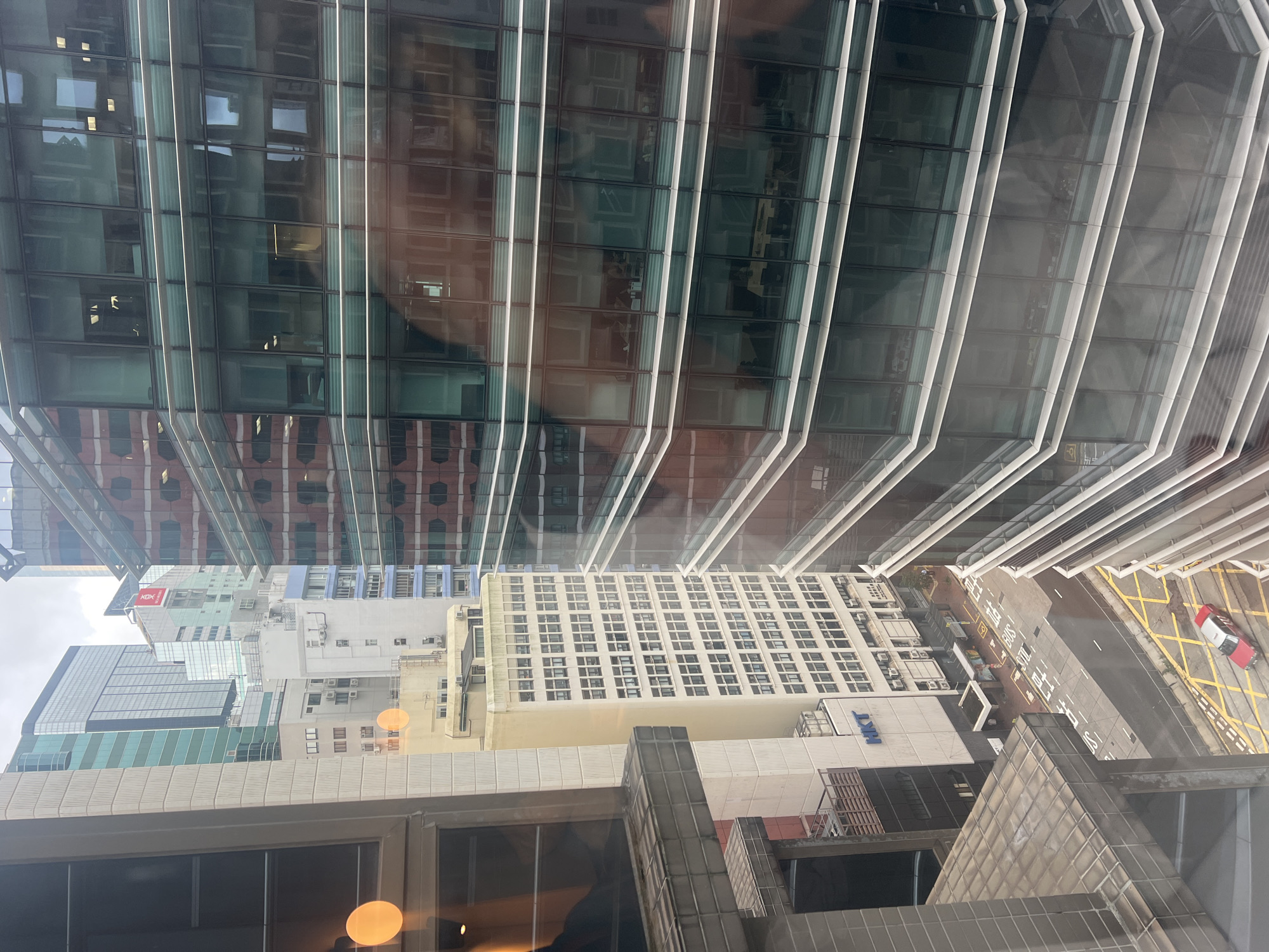This image captures a street view in Hong Kong, China, from a high vantage point, likely an office window. The scene is dominated by modern high-rise buildings with reflective glass facades, showcasing the dense urban landscape characteristic of the city. In the foreground, a large building with a white exterior and numerous windows is partially visible on the left. Adjacent to it is a taller building with a striking glass exterior featuring horizontal white banding. To the right, another building displays a red and glass facade, reflecting the surrounding structures.
Below, a street is visible with painted yellow and white markings, indicating traffic lanes and pedestrian crossings. A red and white taxi is parked on the street, alongside what appears to be a bus stop. Several smaller structures, possibly shops or kiosks, are located along the sidewalk. The overall atmosphere is one of a bustling urban environment, with a clear sense of verticality due to the towering skyscrapers. The lighting suggests it is daytime, with overcast conditions indicated by the diffused light and the subtle reflections on the glass surfaces.