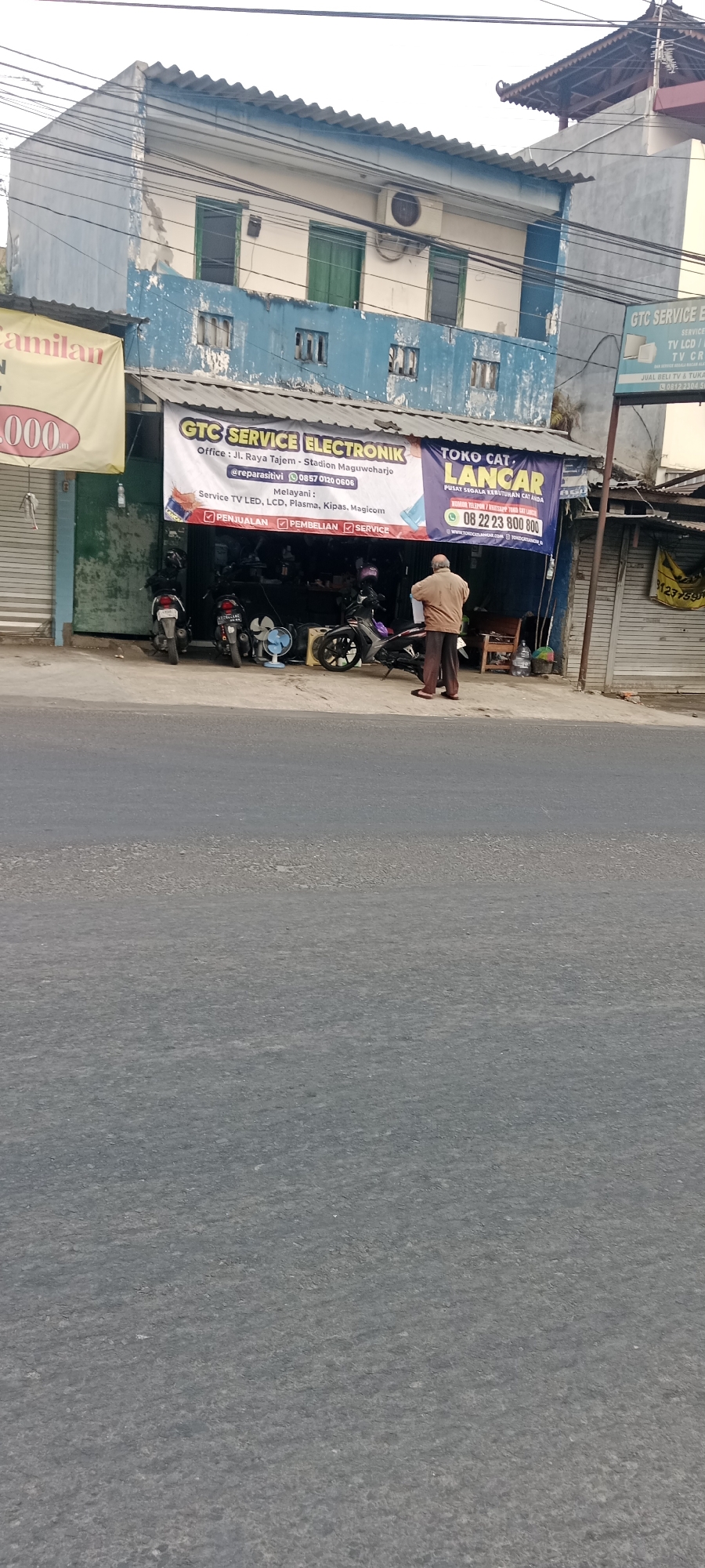 The image shows the exterior of a storefront in Maguwoharjo, Indonesia. The building is a two-story structure with a blue facade, showing signs of wear and peeling paint. Above the entrance, a banner displays "GTC SERVICE ELEKTRONIK" and lists services such as TV LED, LCD, Plasma, Kipas, and Magicom, along with contact information. To the right, another banner advertises "TOKO CAT LANCAR" (Lancar Paint Store). Several motorcycles are parked in front of the storefront. A man in a light brown shirt and dark pants is standing outside the store, facing away from the camera, holding what appears to be a piece of paper. The street in front of the building is paved with asphalt. Electrical wires are visible overhead.