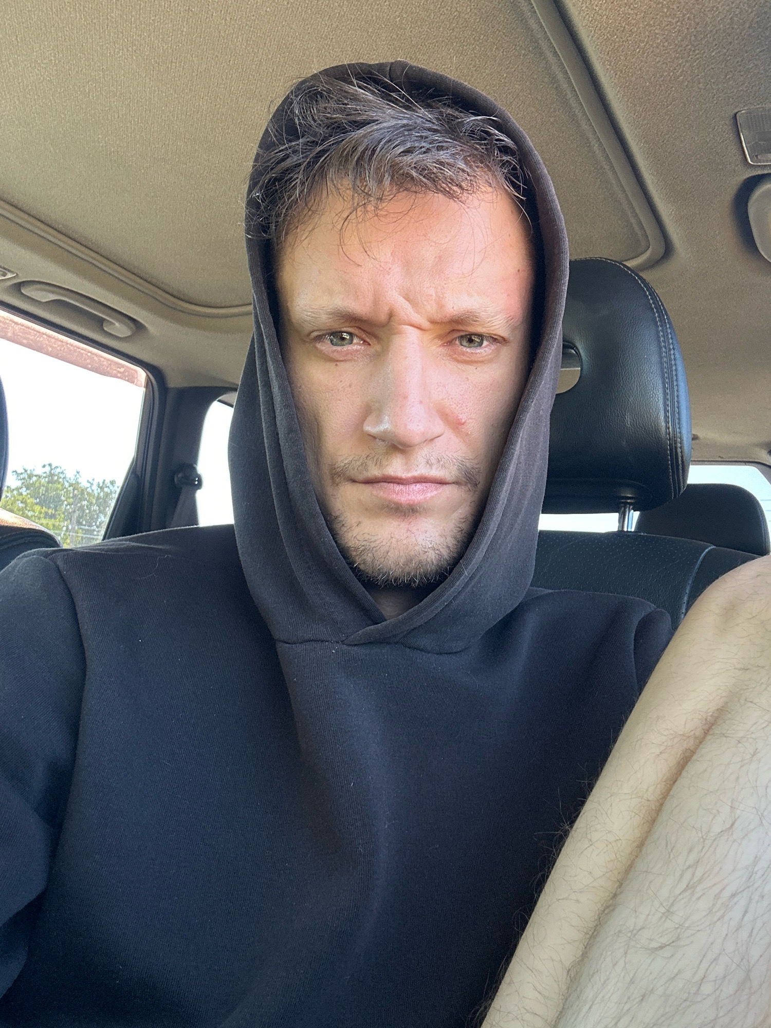 A man wearing a black hooded sweatshirt sits in the driver's seat of a car, looking directly at the camera with a furrowed brow and a slight grimace. The overhead interior of the car, including the sunroof, is visible. Outside the car window, trees and foliage are present. The man has light brown hair with streaks of gray and stubble on his chin and cheeks. His right arm and leg are visible to the right of the frame, covered in light-colored hair. The image was taken in Makiv, Ukraine.